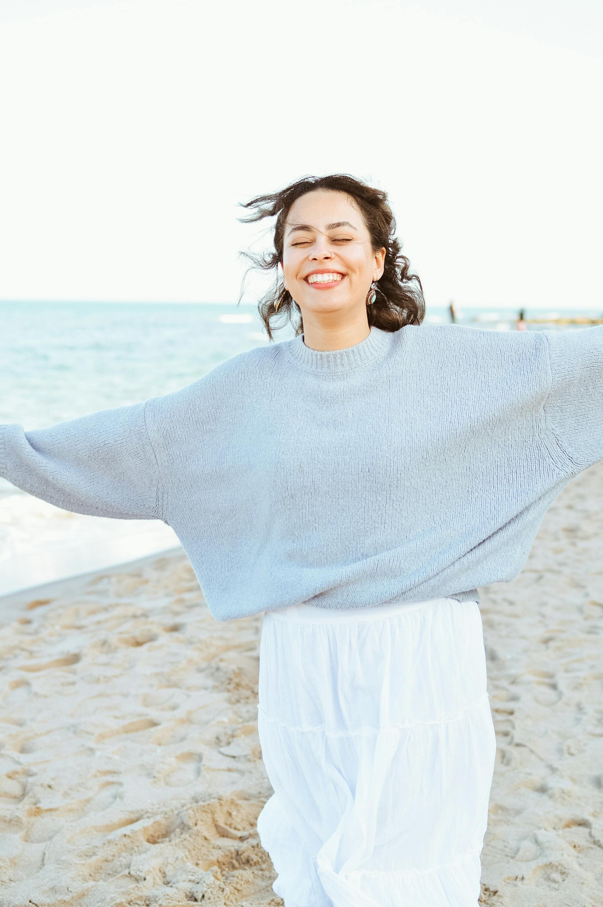 Smiling woman with arms wide open on a sunny beach day, embracing the moment.
