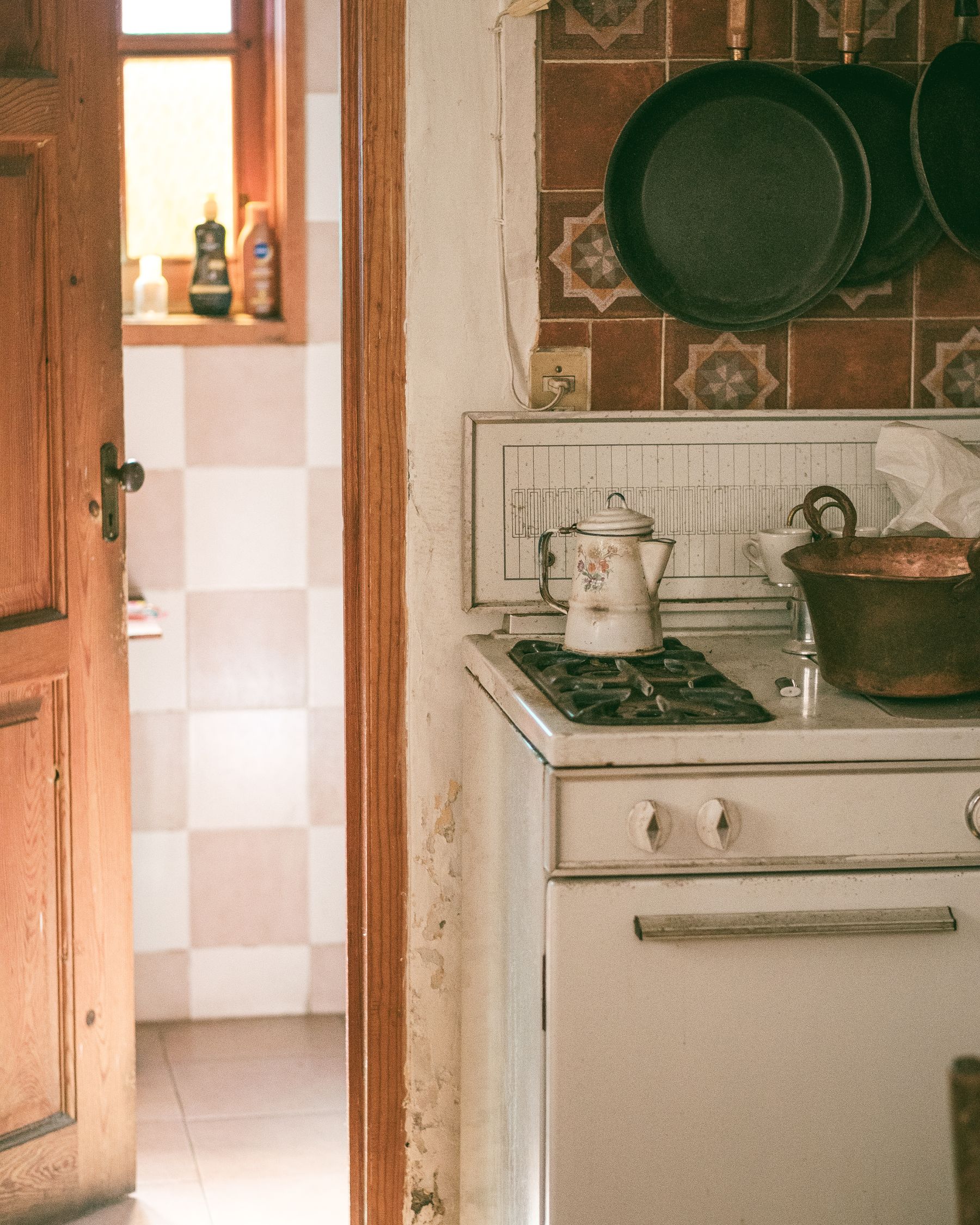 Older white cooker beside a wood door, patterned brown tiles and hanging pans