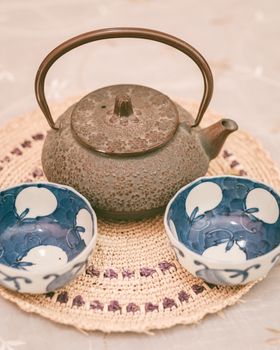 A rustic cast iron teapot accompanied by two decorated blue and white ceramic cups on a woven mat.