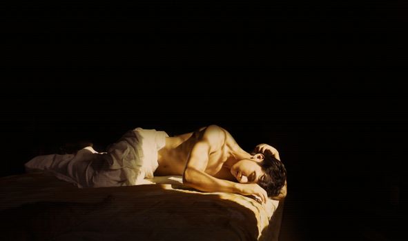 A dramatic artistic portrait of a nude man reclining in low light with a contemplative pose.