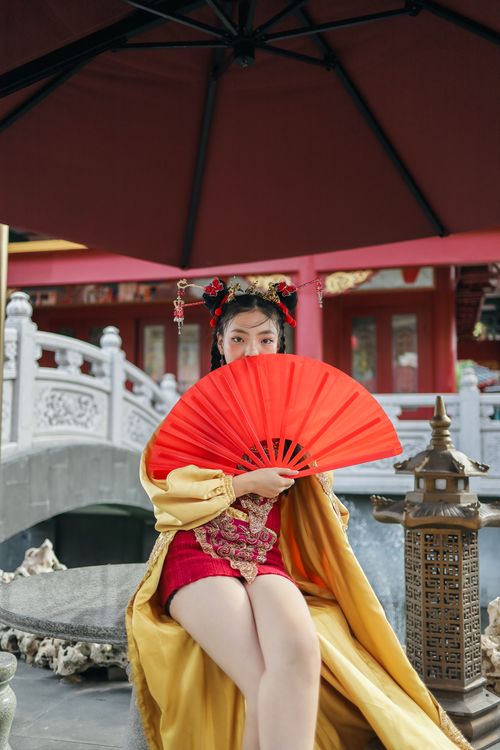Free Woman in traditional Chinese dress sitting by a bridge, holding a vibrant red fan. Stock Photo