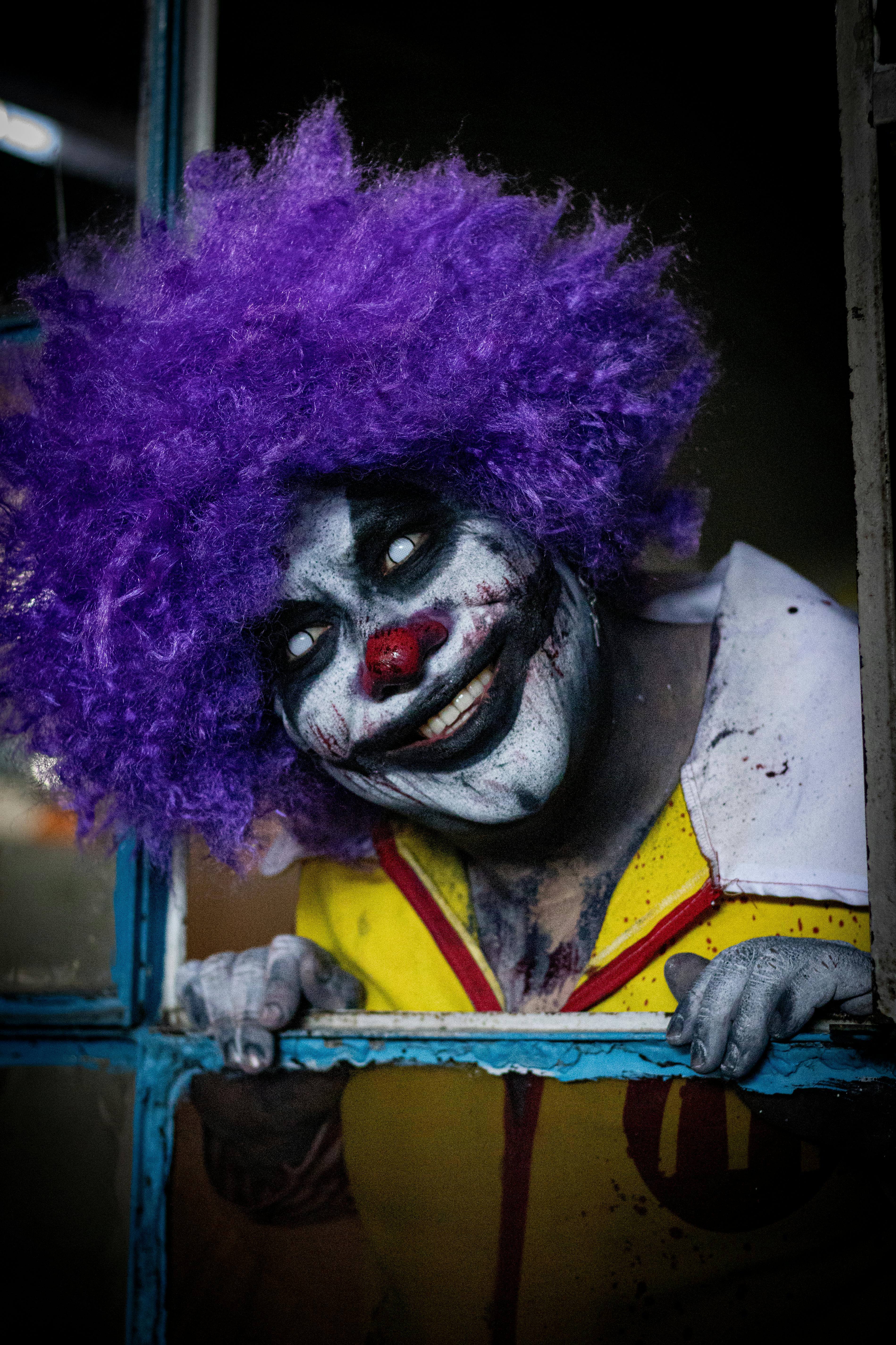 A creepy clown with purple wig and haunting makeup peering through a window, perfect for Halloween themes.