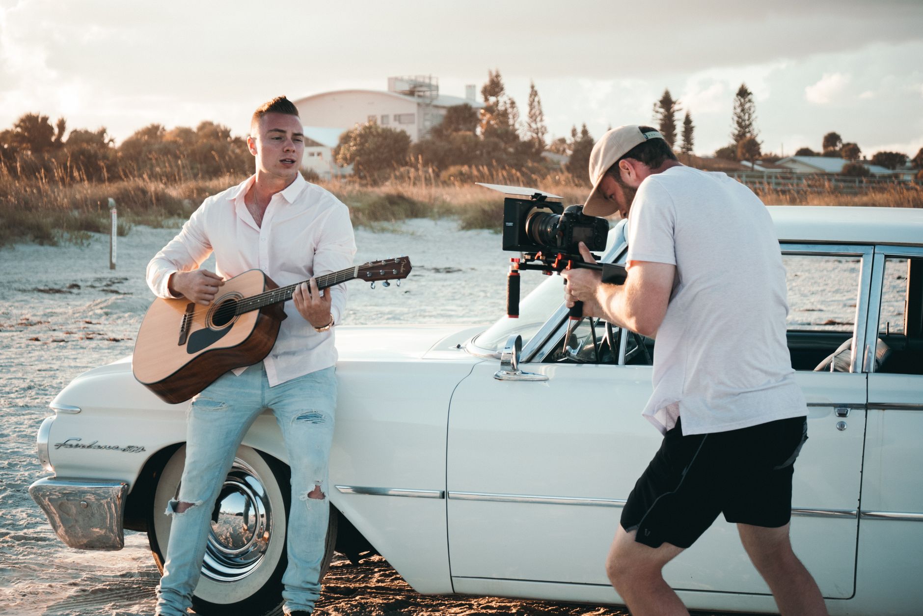 A young man plays guitar as a videographer films by a vintage white car on a sunny beach day.