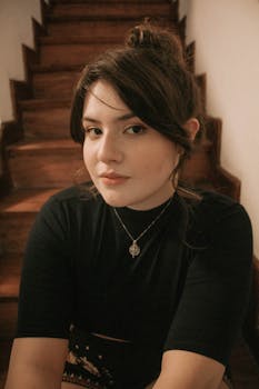 A young woman sitting on wooden stairs, thoughtfully posing indoors.