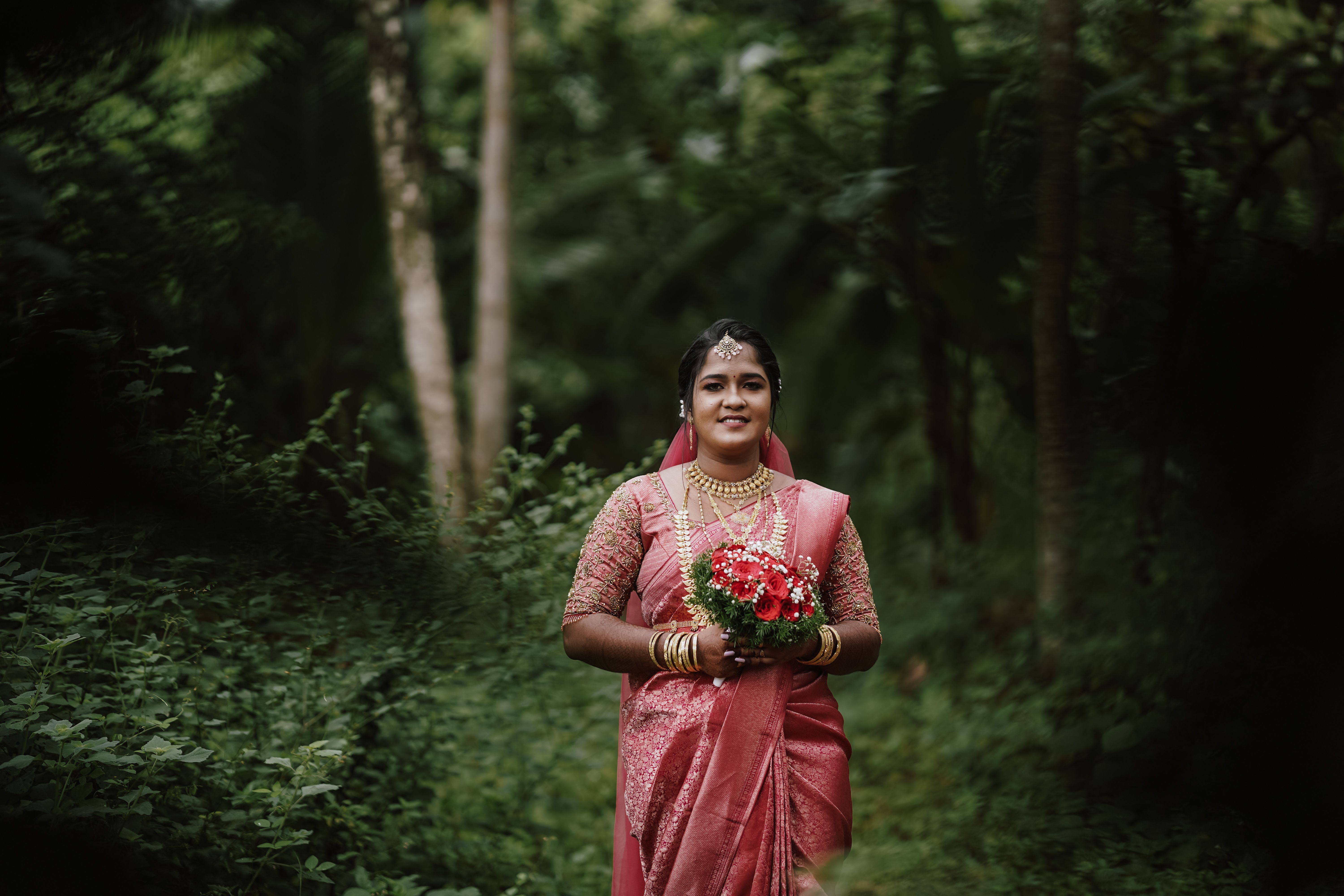 South Asian bride in pink saree holding bouquet in verdant Chennai forest setting.