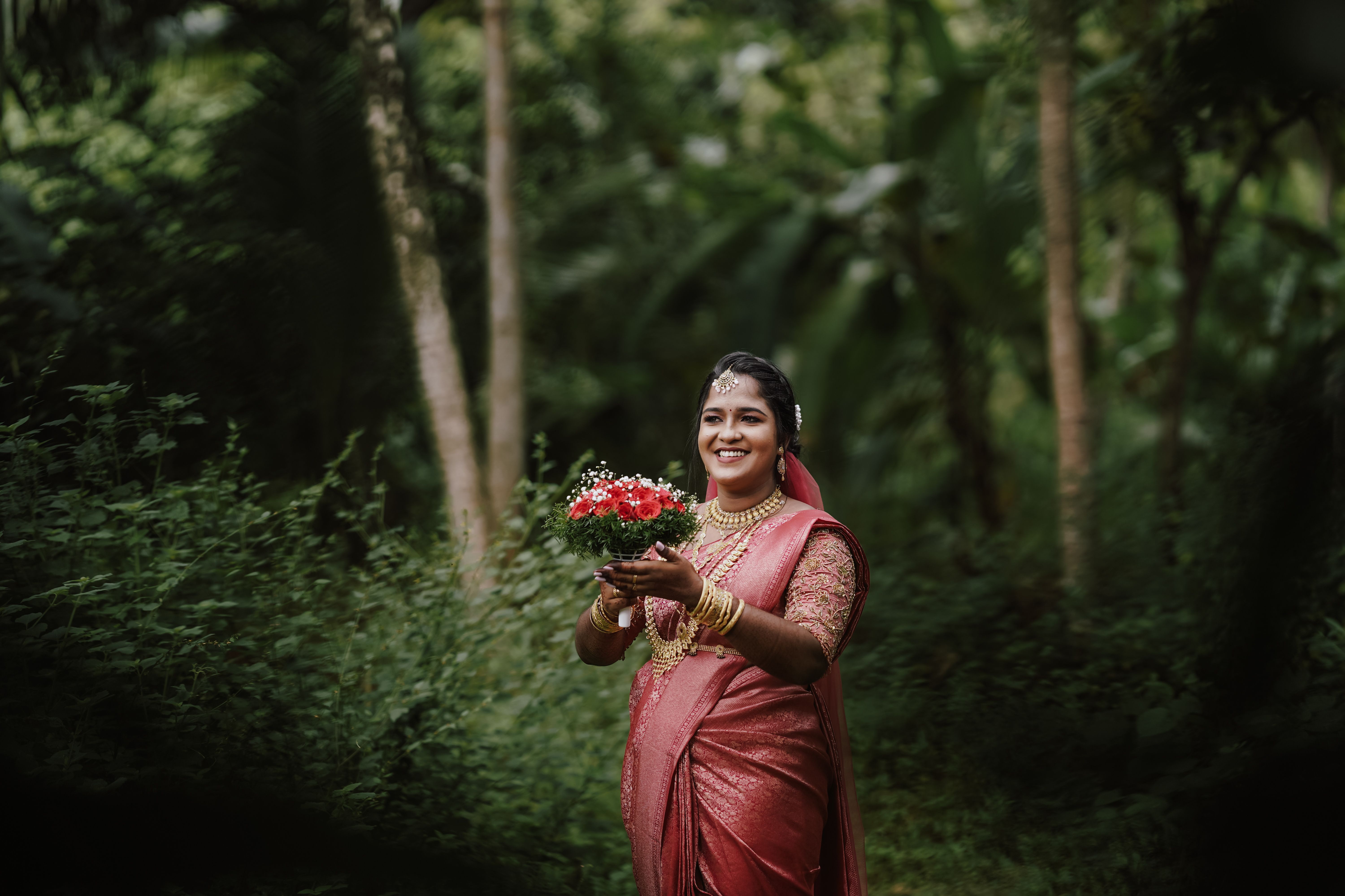 A happy bride in a traditional saree holding flowers in a lush outdoor setting.