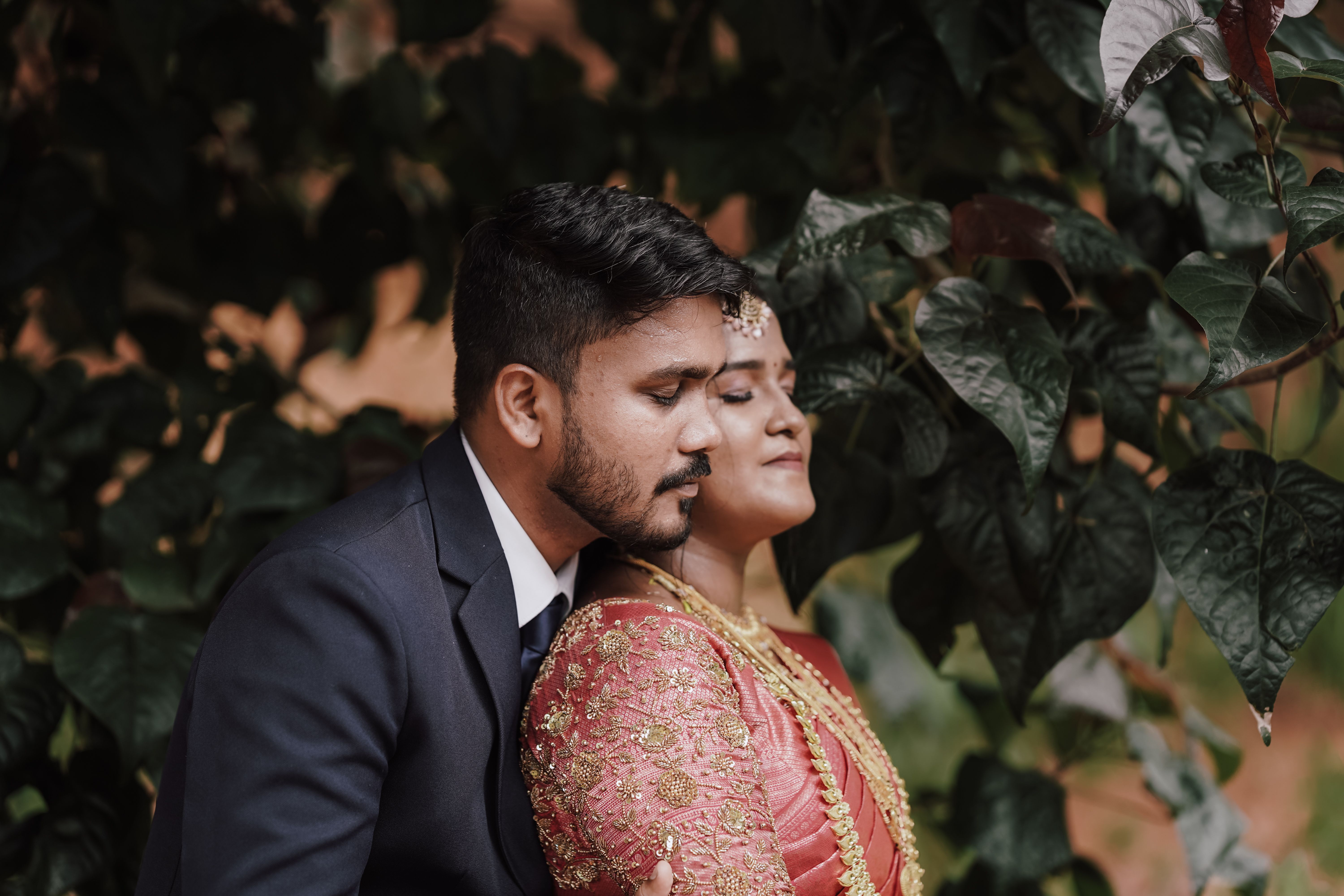 A South Asian couple in traditional attire shares a romantic moment amidst lush greenery.