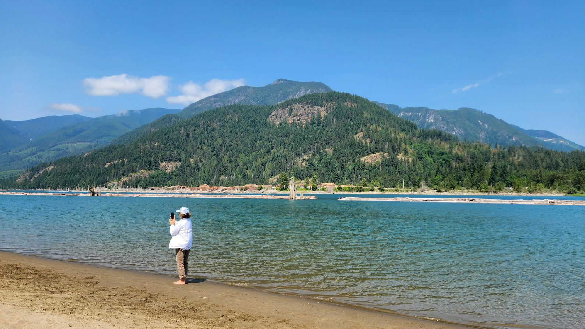 A person photographing a river and mountainous landscape during a sunny day.