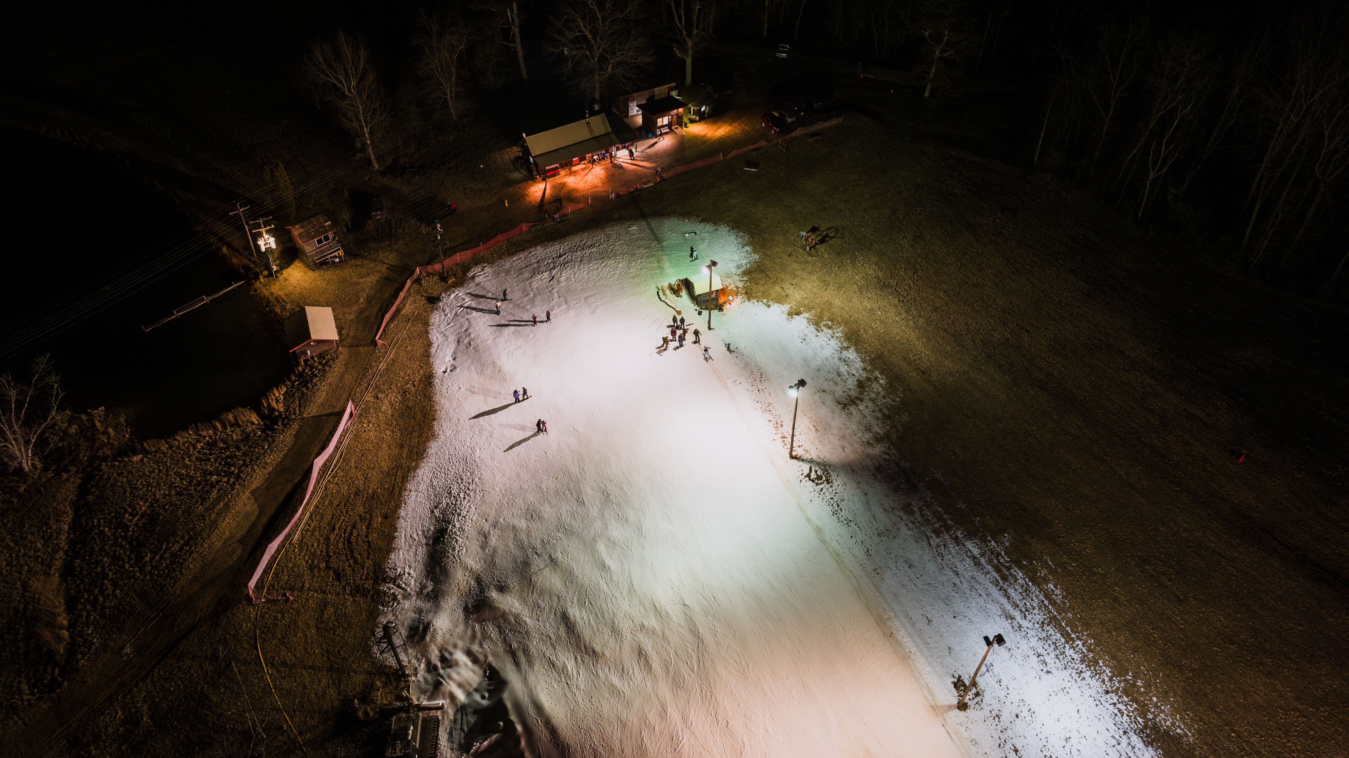 Aerial shot of a night-time ski slope with artificial snow, illuminated by lights and small crowd gathered.