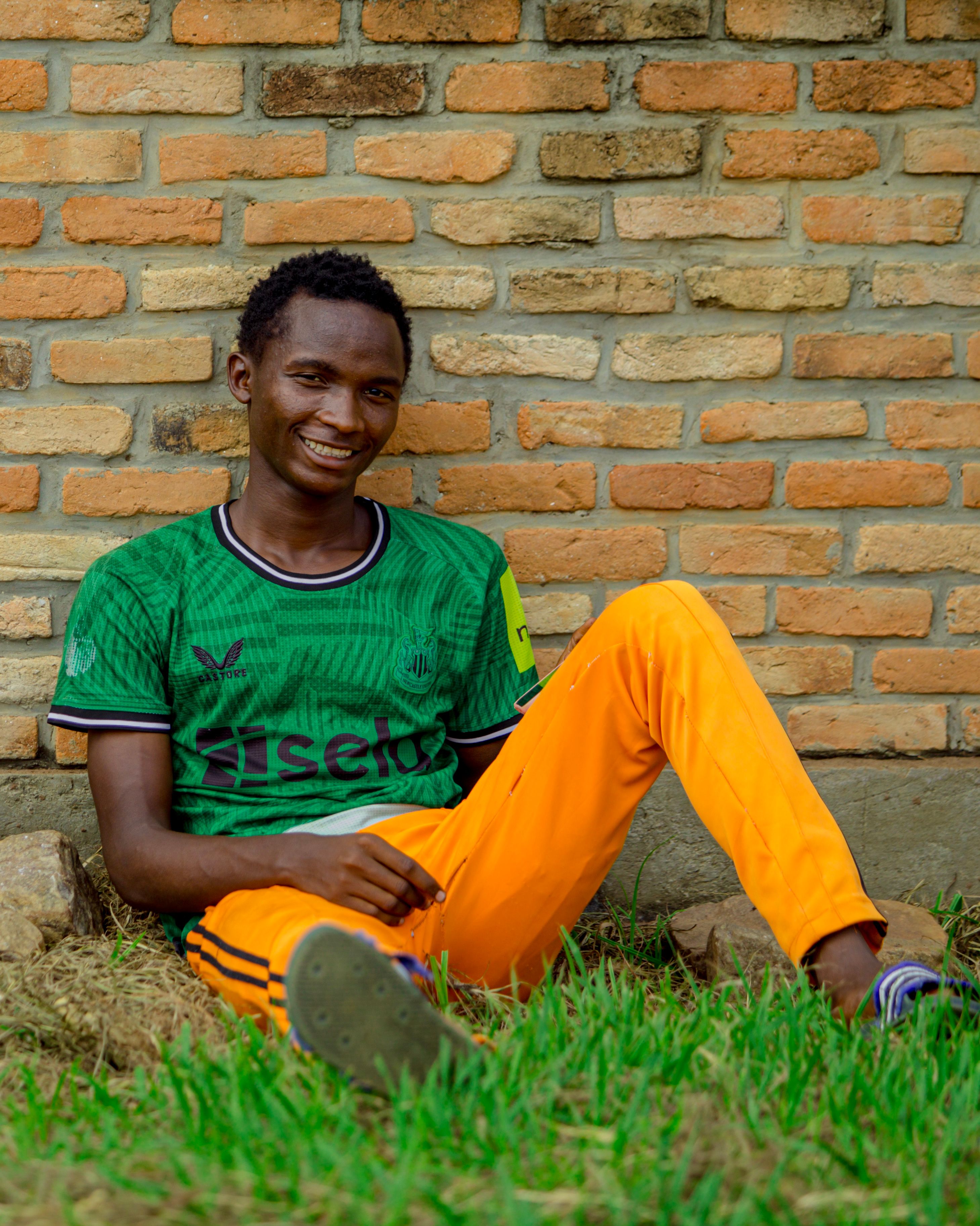 A young man in sports attire sits against a brick wall, smiling and relaxed.