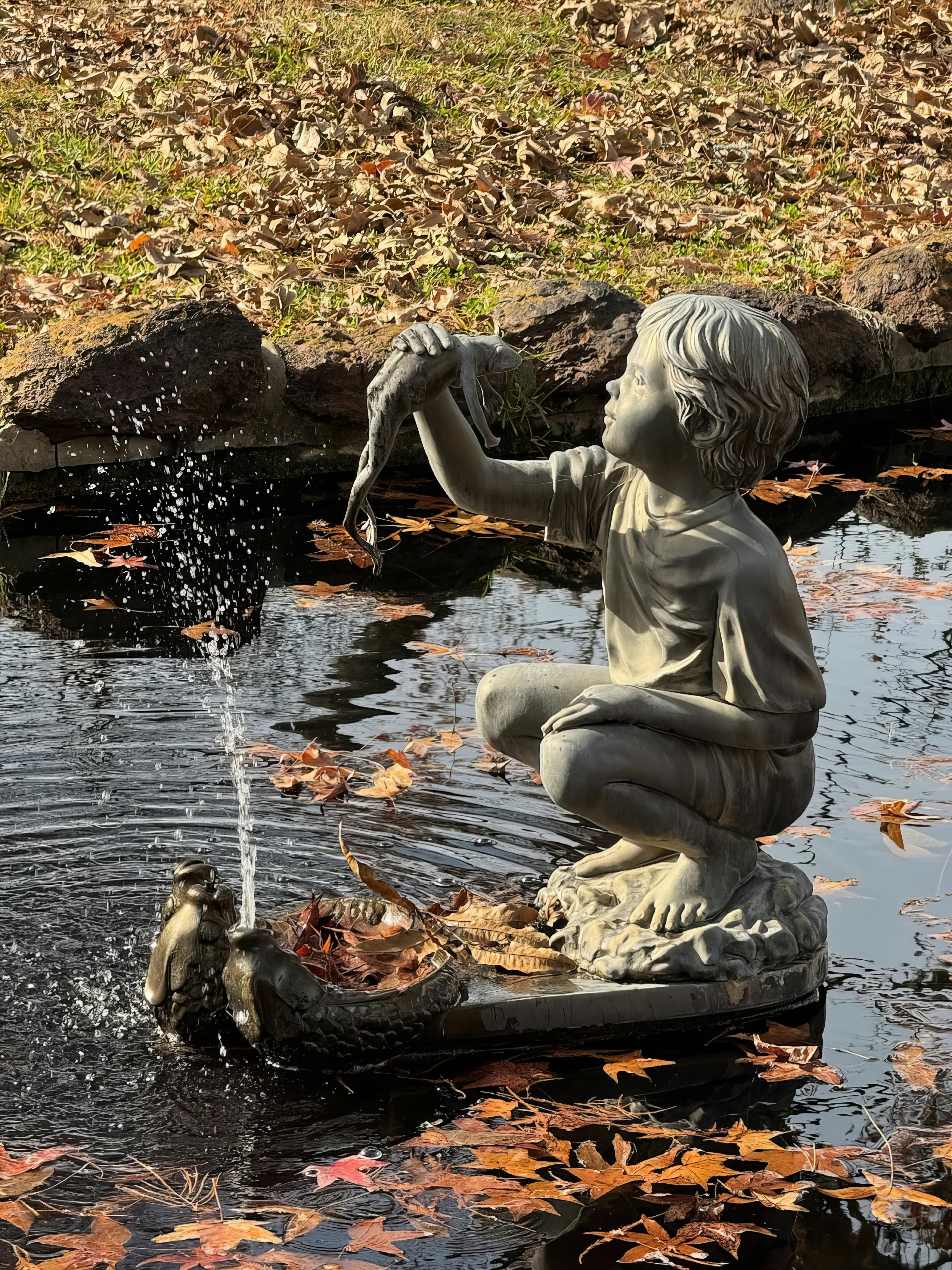 A stone statue of a boy holding a fish in a serene pond surrounded by autumn leaves.