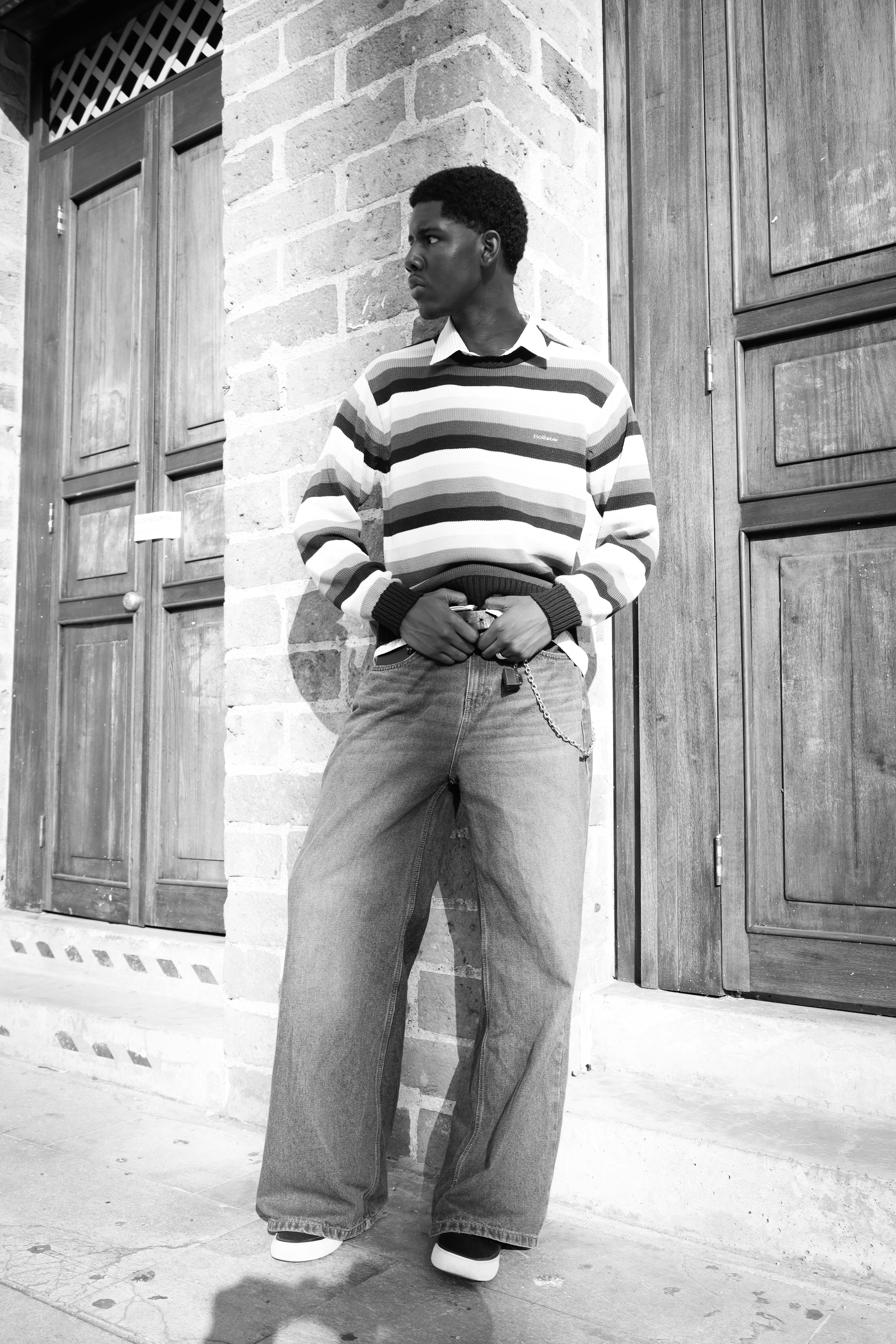 Black and white photo of young man in casual outfit standing outdoors by a brick wall.
