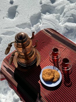 Copper teapot and glasses of tea on snowy outdoor setup with cookies.