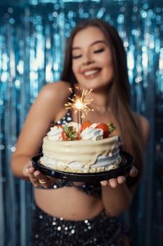 A happy young woman holds a cake with a sparkler against a festive backdrop, celebrating a birthday in Brazil.