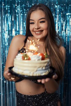 A happy woman holding a birthday cake with a lit sparkler against a festive background.