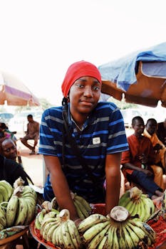 A female vendor in Abuja, Nigeria selling bananas at a lively street market under umbrellas.