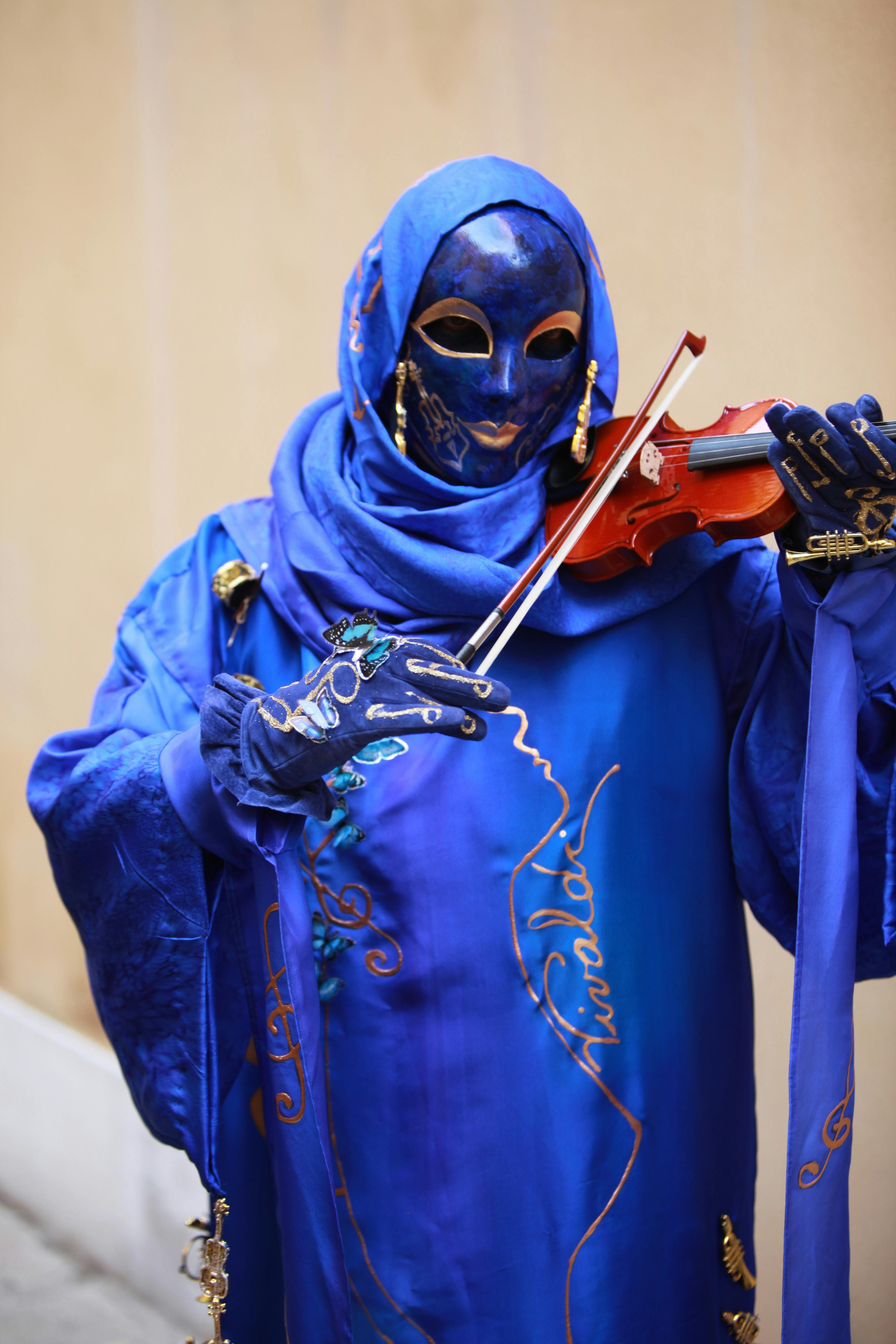 Ücretsiz Canlı mavi cübbeli maskeli sanatçı keman çalarak sokak ortamına gizem ve sanat katıyor. Stok Fotoğraflar