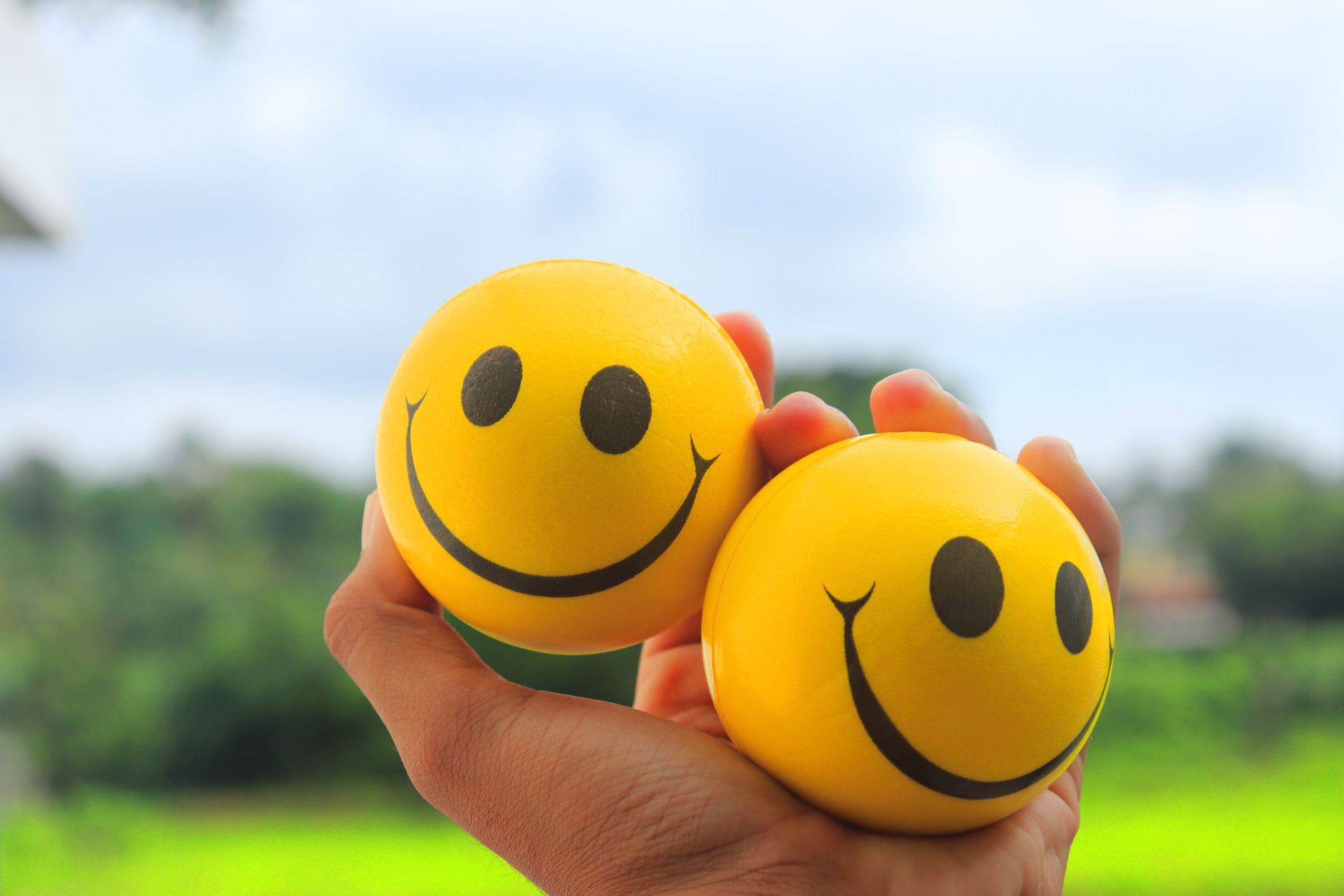 Two yellow smiley face balls in a hand against a vibrant outdoor background.