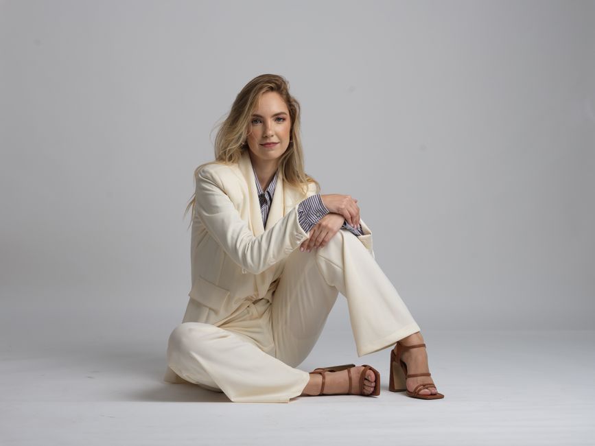 Elegant woman seated in white suit on plain background, exuding confidence.