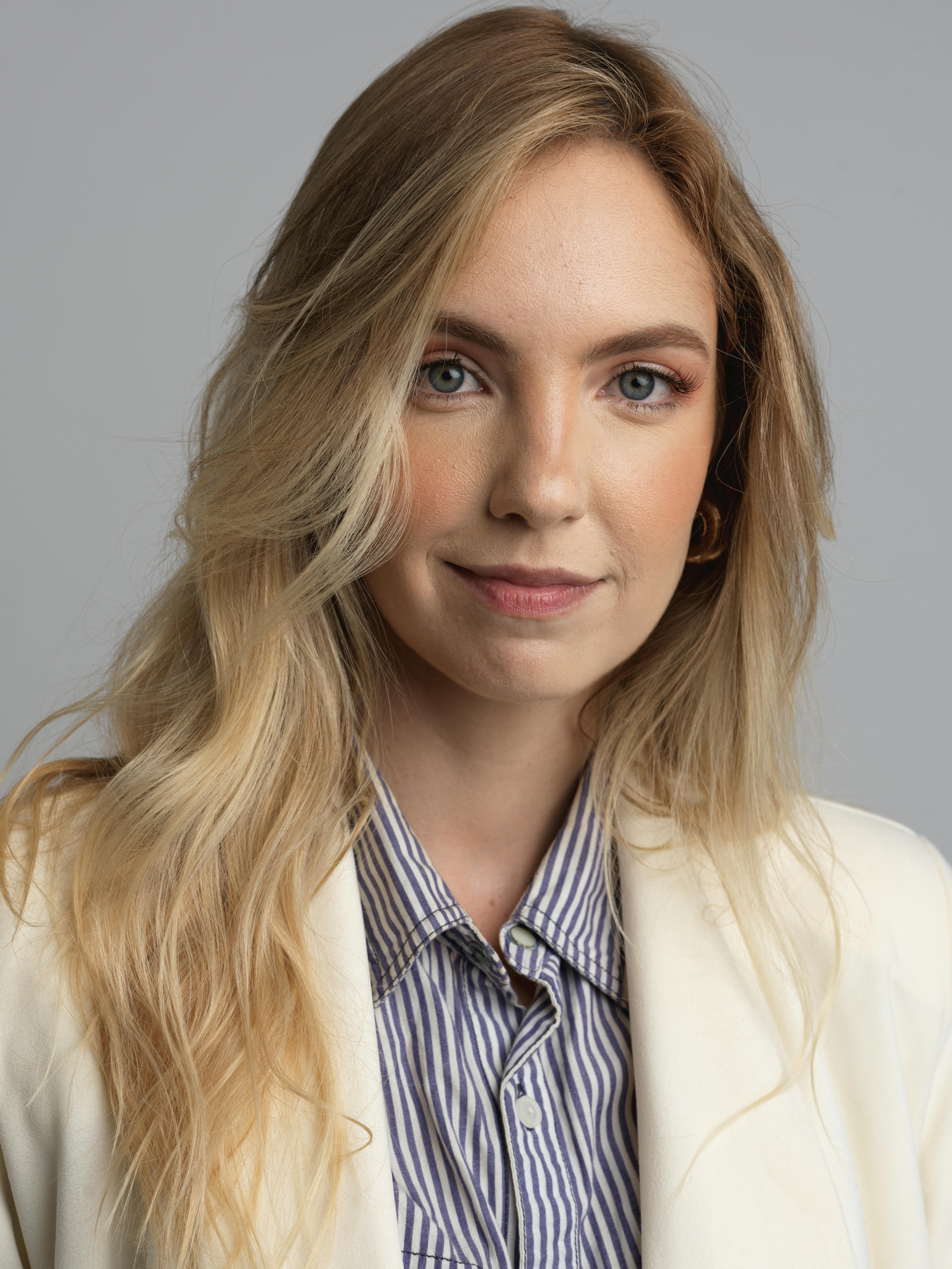 Portrait of a young professional woman in a striped shirt and white blazer, with long blonde hair.