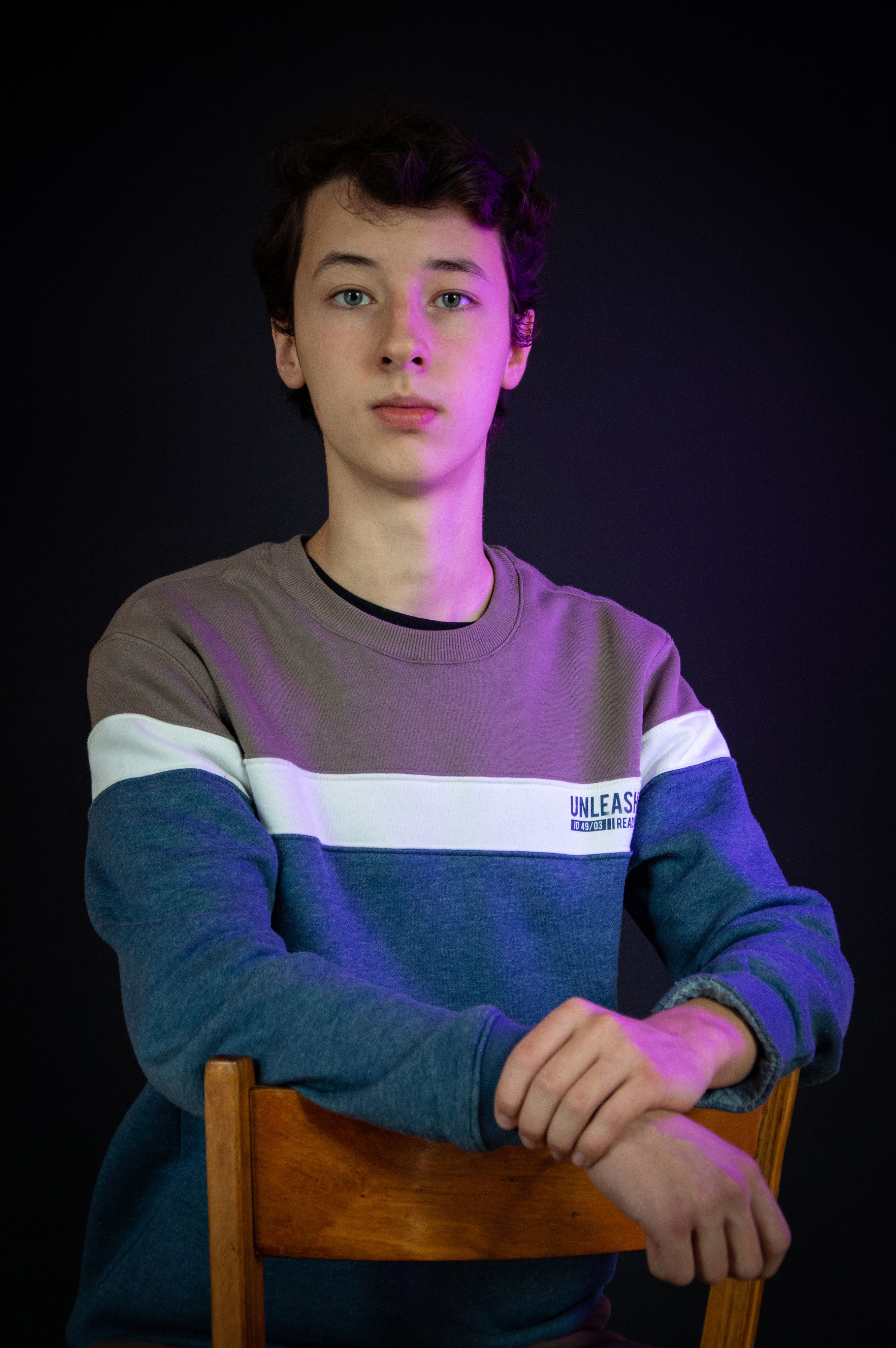 Studio portrait of a young man sitting on a chair with dramatic lighting and dark background.