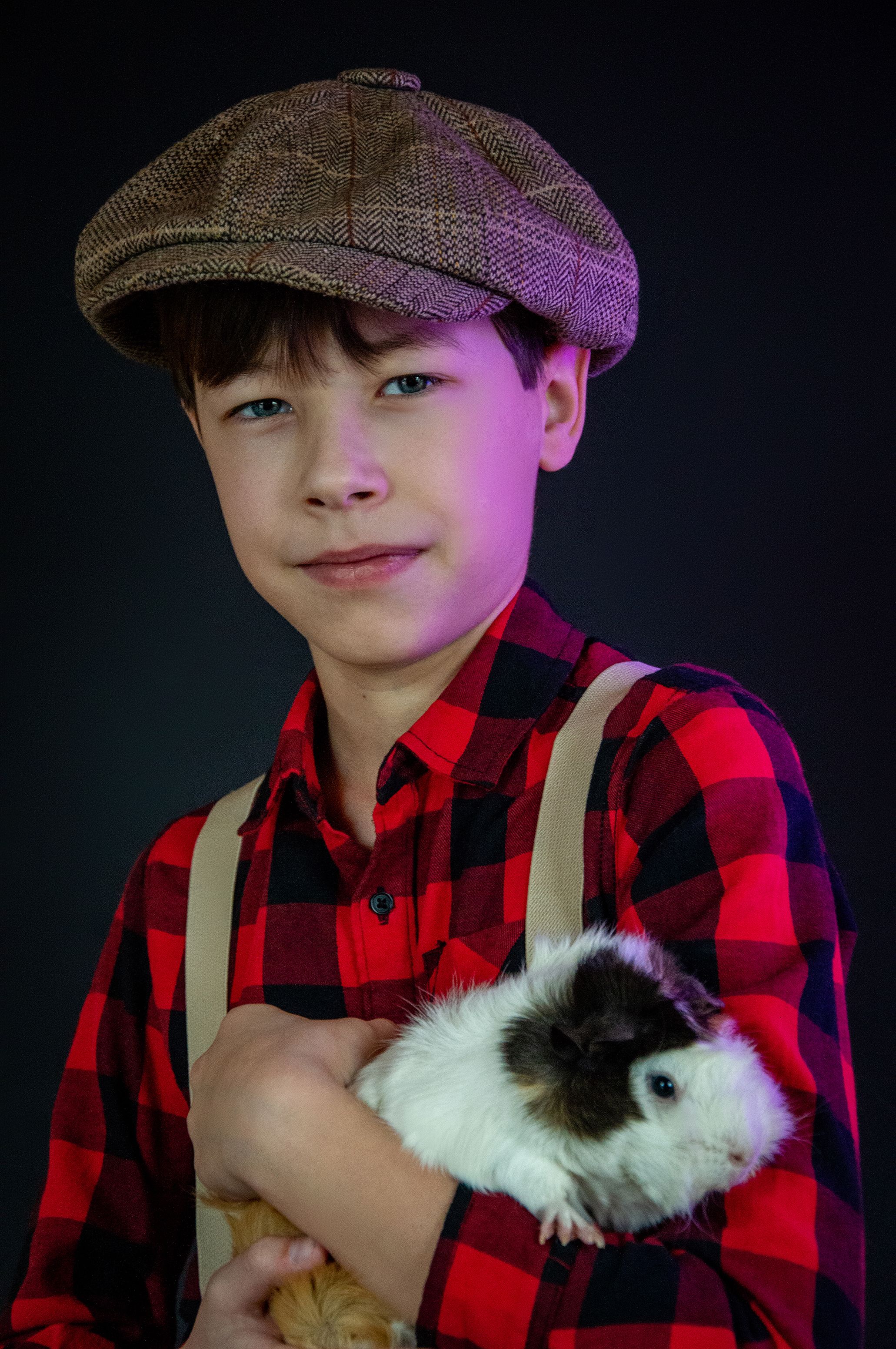A young boy in a checkered shirt and cap holding a guinea pig, studio portrait.