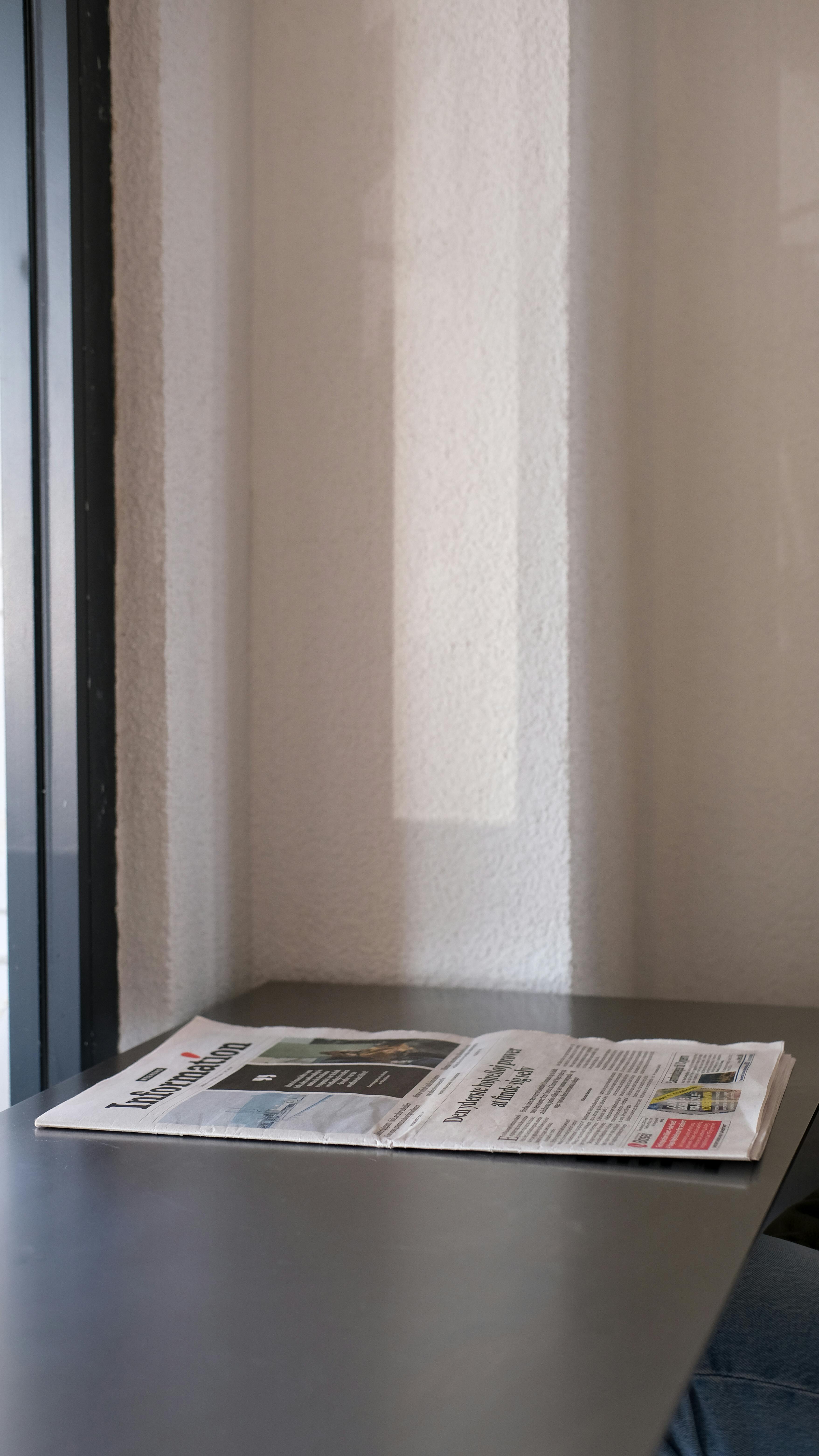 Gratis En brettet avis hviler på et bord nær et vindu i minimalistisk innendørs setting. Arkivbilde