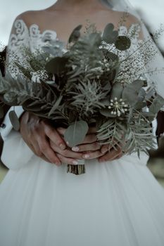 Bride and groom holding a natural bouquet on their wedding day. Elegant and romantic scene with greenery.