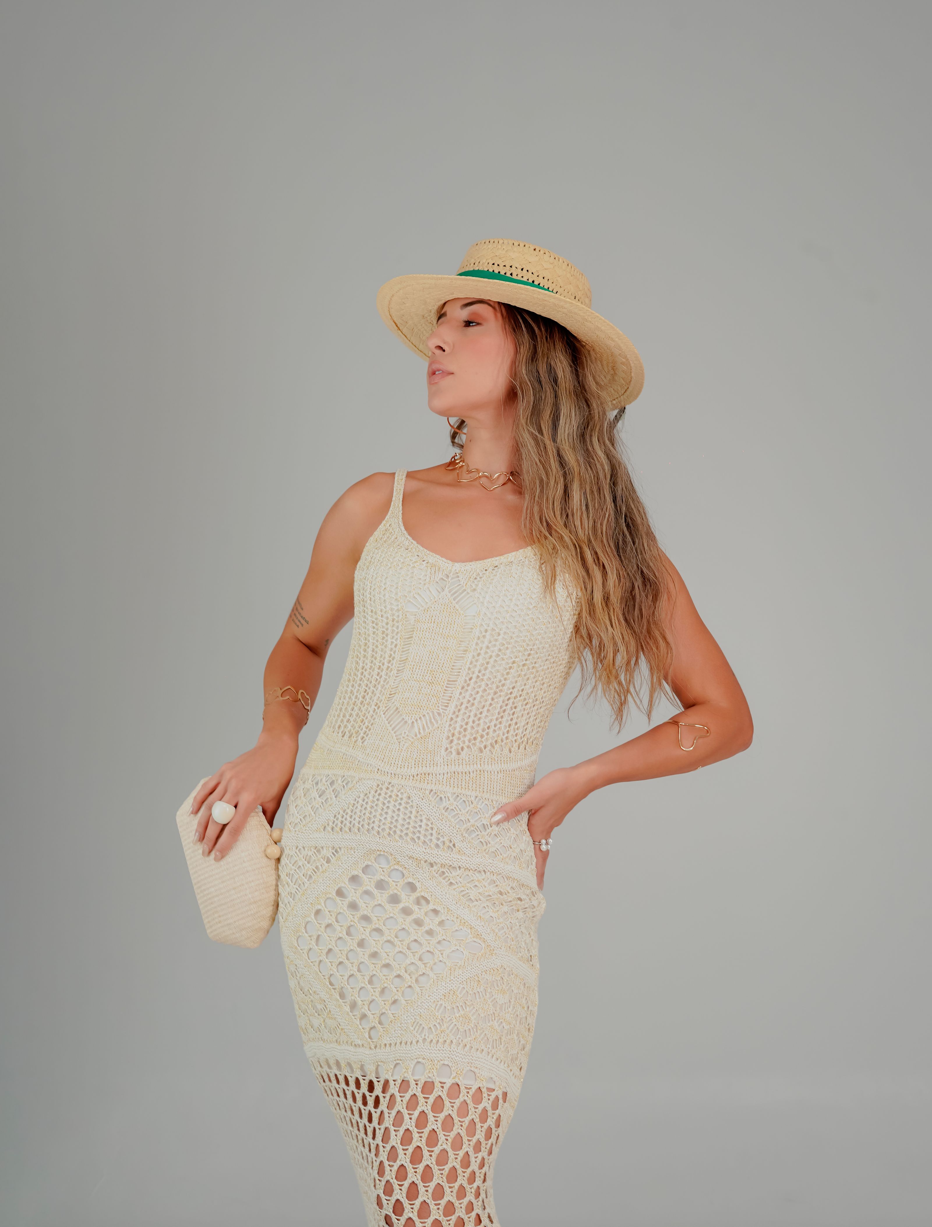 Mulher estilosa com um vestido de crochê e chapéu de palha posando confiante em um fundo neutro.