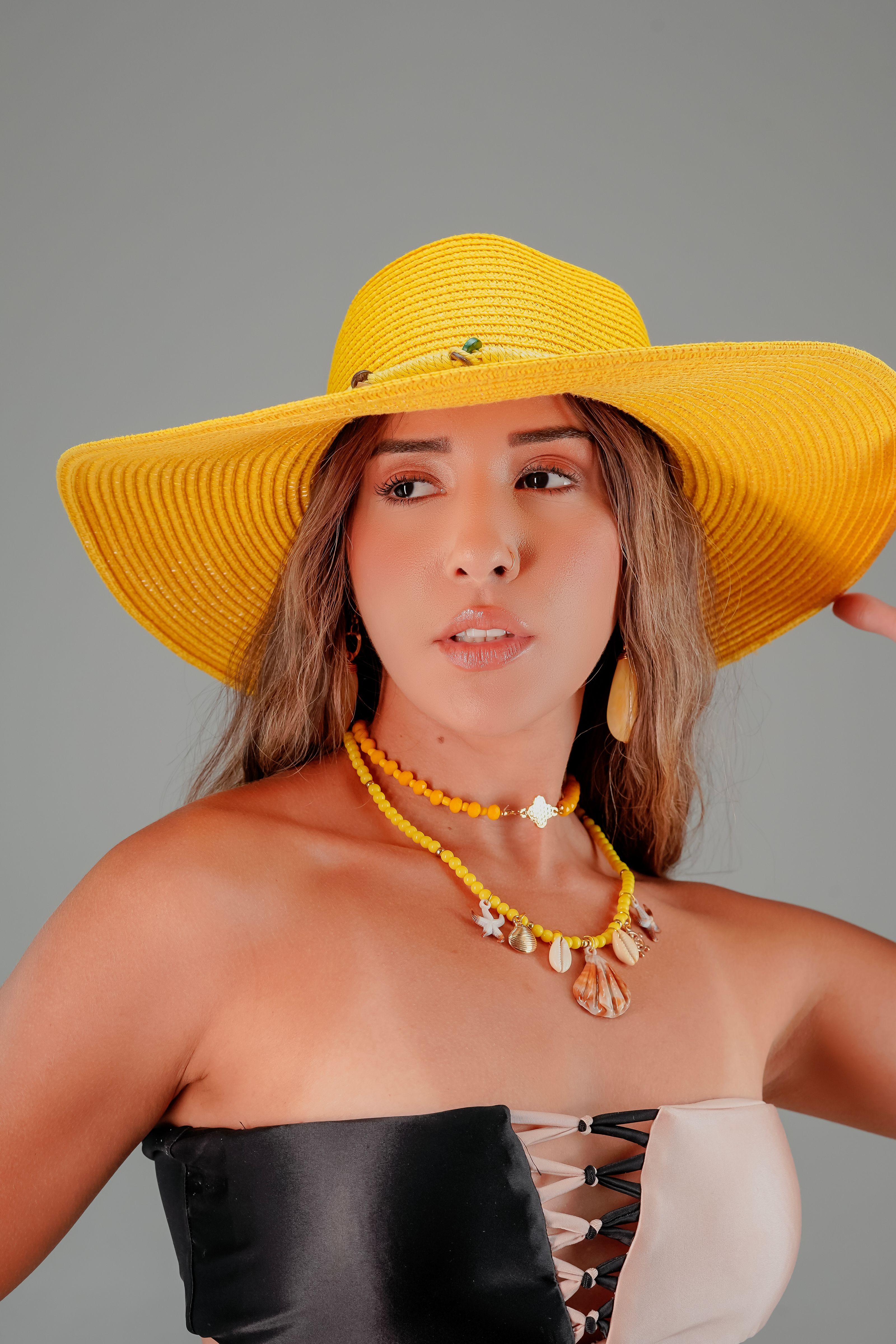 Retrato de uma mulher estilosa usando um chapéu de sol amarelo e um colar com um fundo neutro.