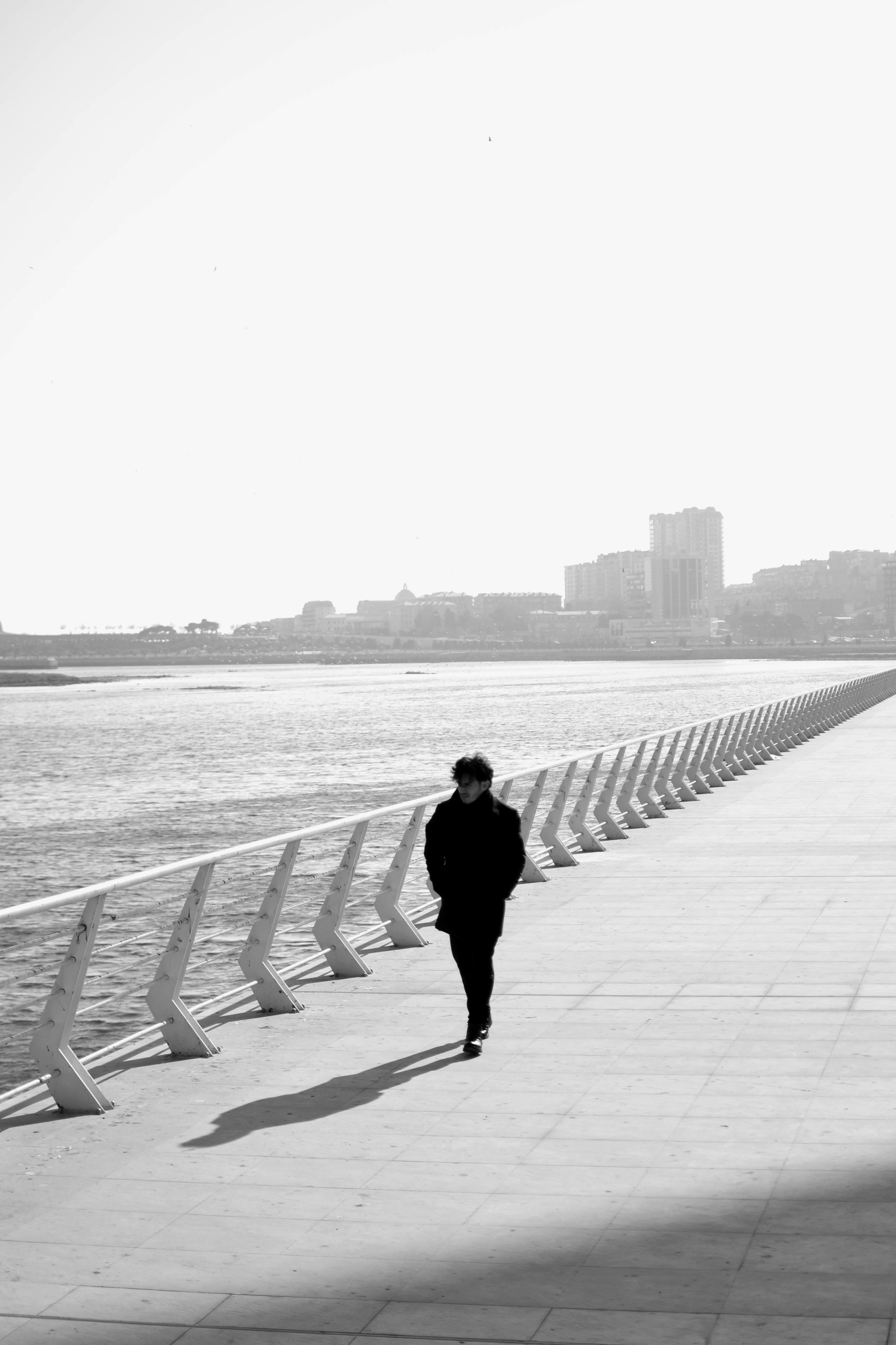 A person walks alone on a seaside promenade with cityscape in stark black and white tones.