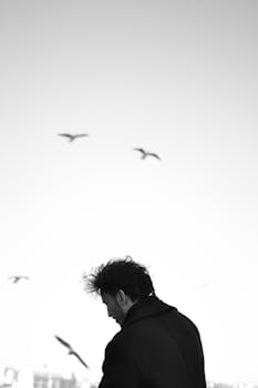 Black and white photo of a man's silhouette with birds flying overhead against a bright sky.