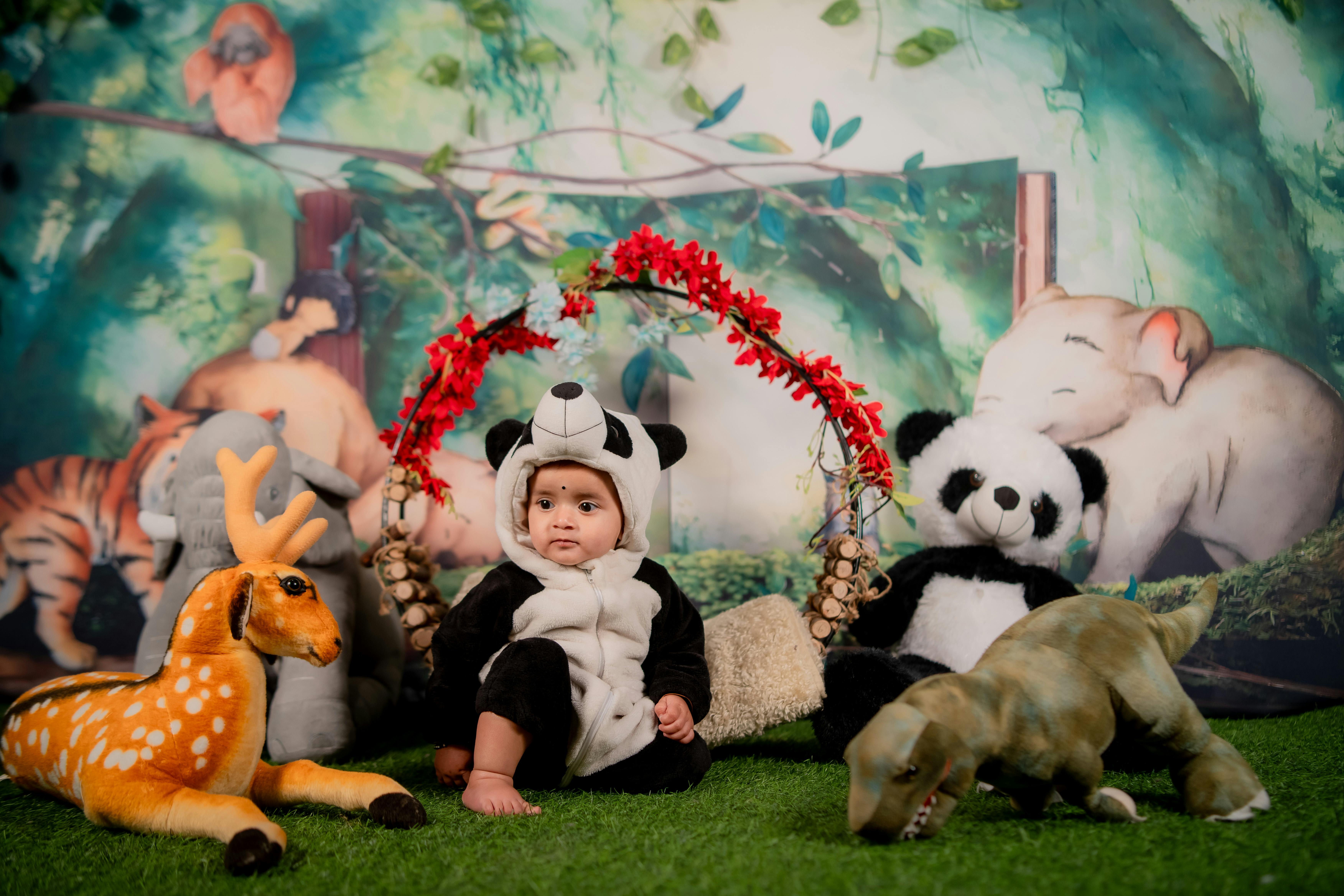 Charming baby in panda outfit surrounded by stuffed animals in a jungle-themed setting.