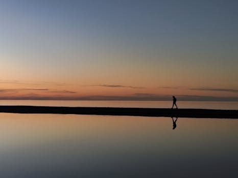 A serene sunset reflection scene with a person walking along the beach, creating a silhouette.