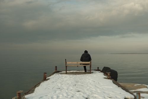 Free Person sitting on a snowy pier bench at Lake Van in Türkiye, reflecting under cloudy skies. Stock Photo