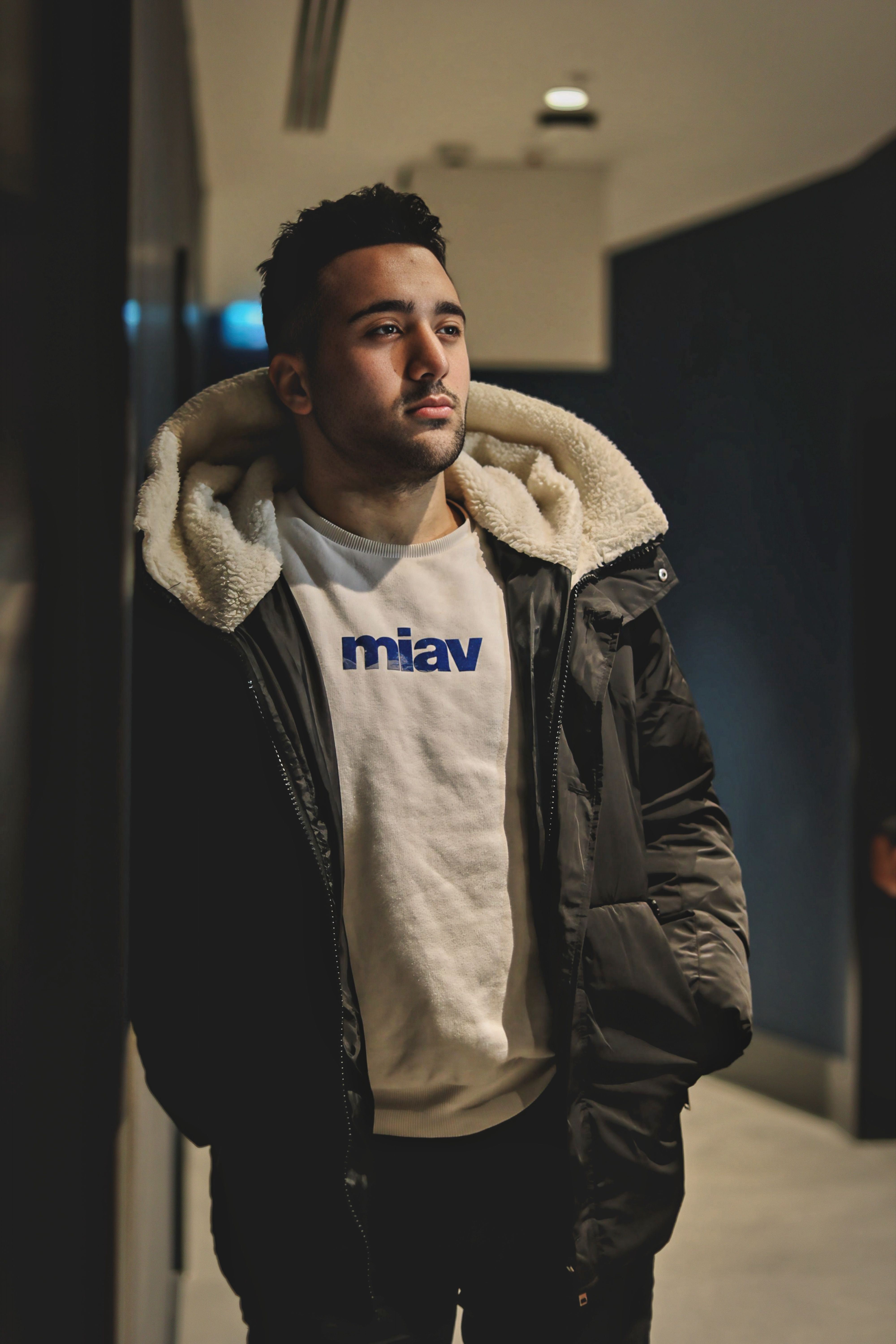 A young man in a winter jacket and hoodie stands indoors, looking thoughtful.