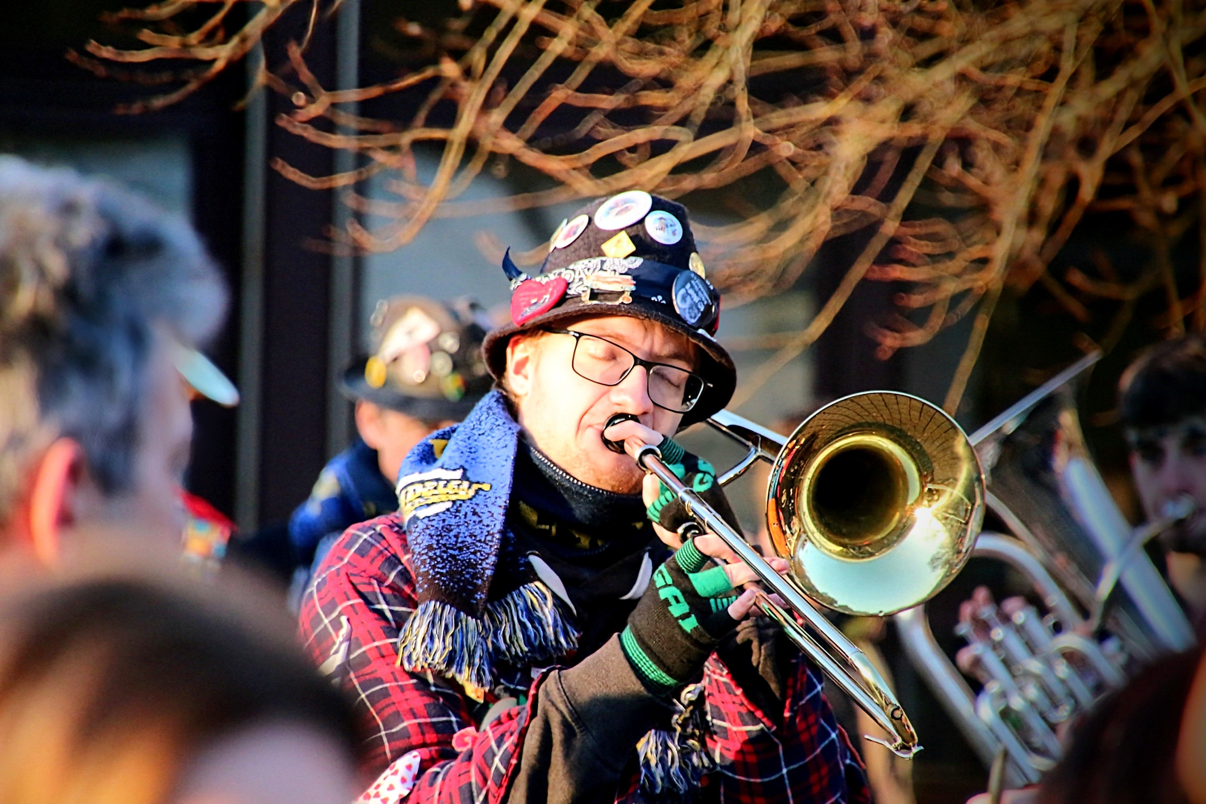 A musician in a plaid jacket plays a trombone during an outdoor performance.