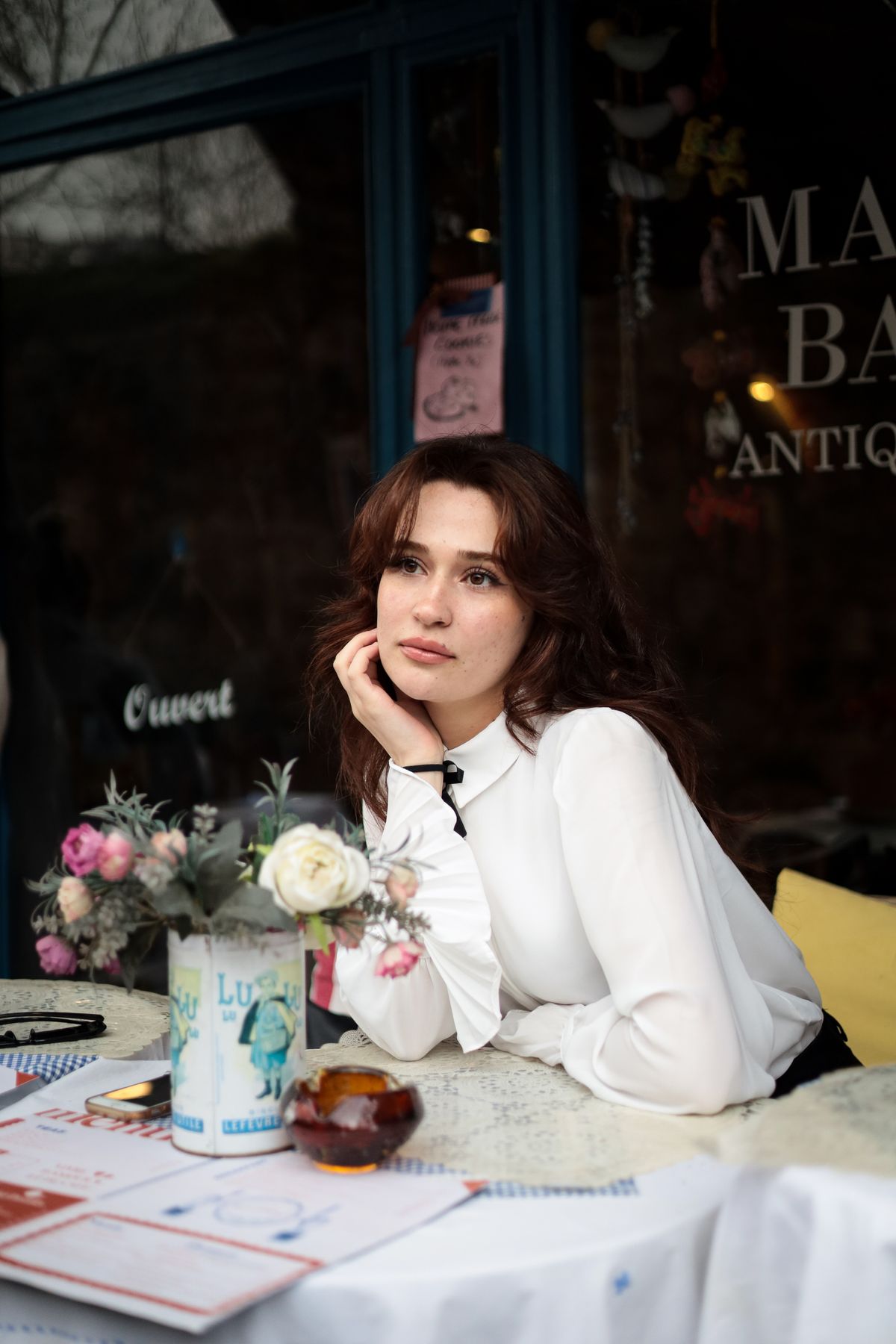 A woman in a white blouse at a cafe table with flowers — effortless Parisian ease
