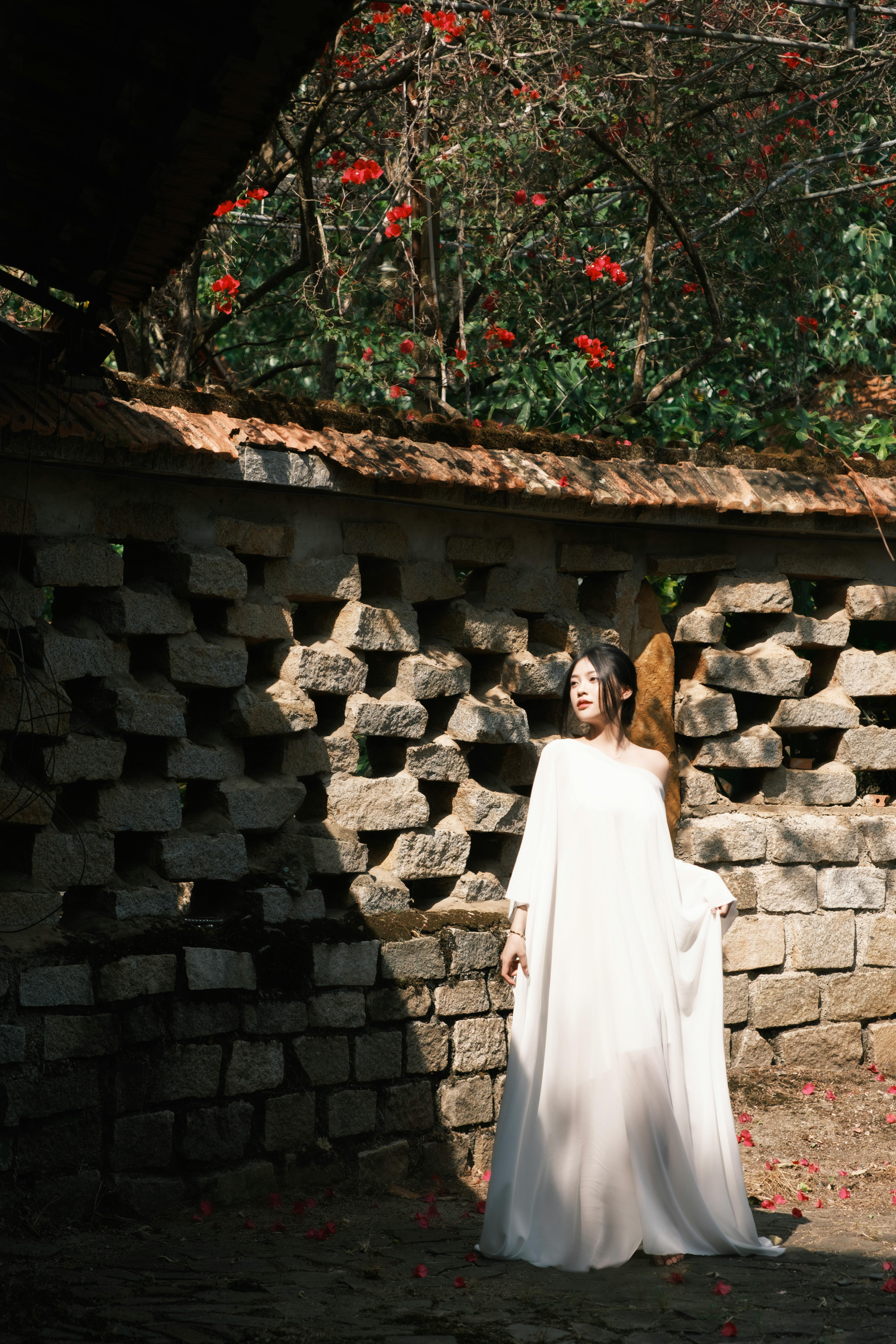 A serene woman stands in sunlight, wearing a flowing white dress, against a stone wall.