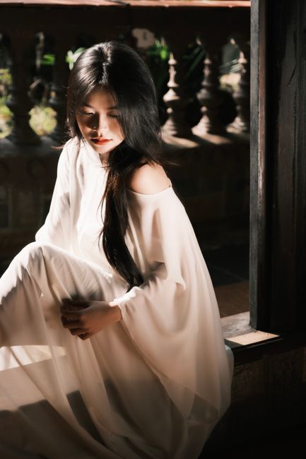 A woman in a flowing white dress sits gracefully in a sunlit, rustic setting.