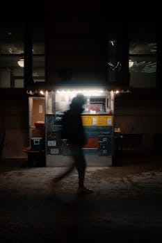Silhouetted person passes by an illuminated street food cart at night.