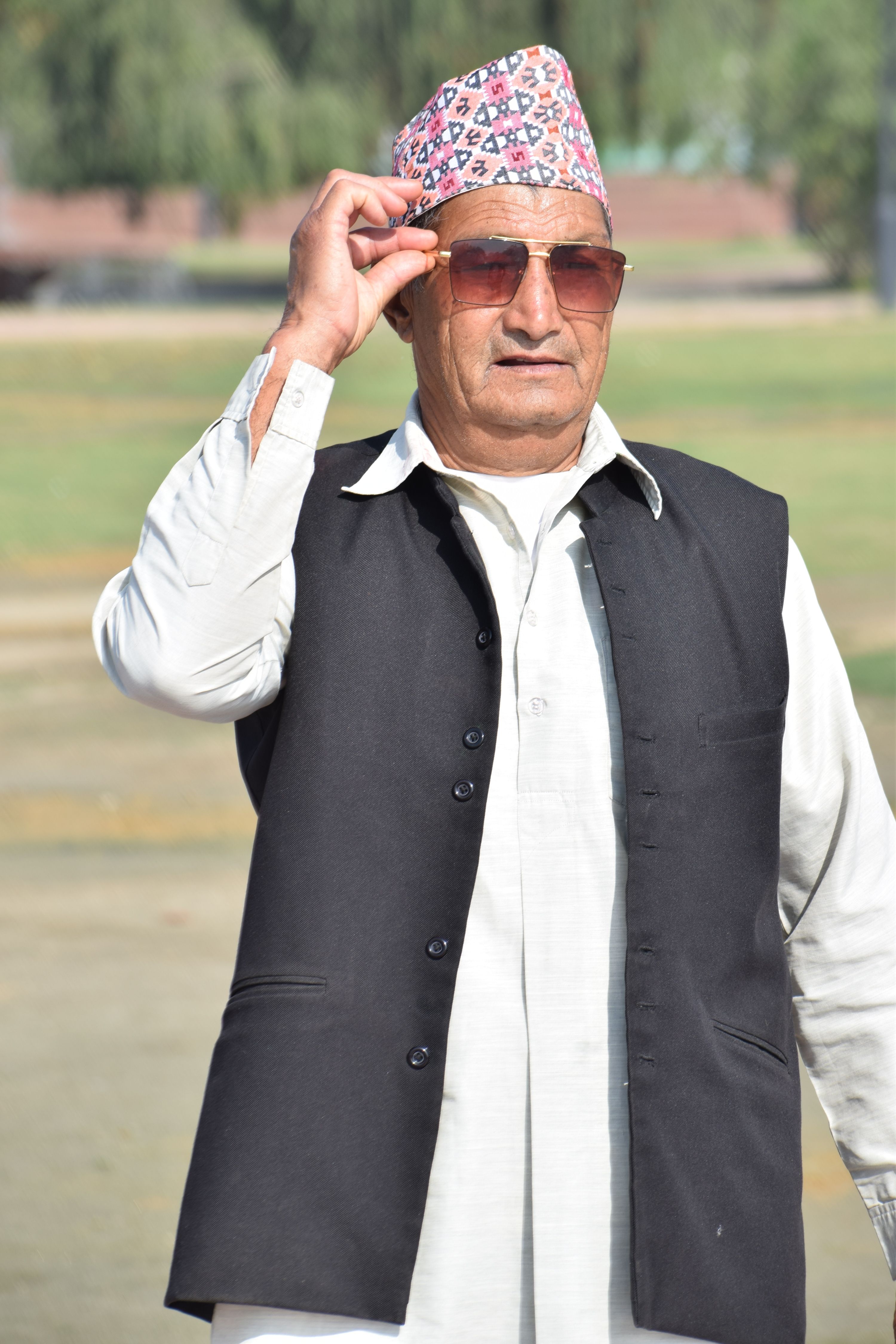 Senior man wearing traditional black vest and hat outdoors, adjusting sunglasses.