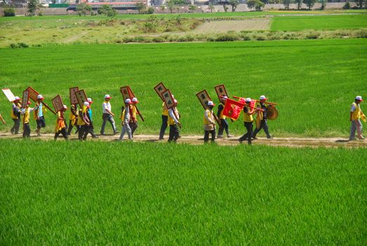 People marching in a line through lush green rice fields holding signs and banners, symbolizing cultural celebration.