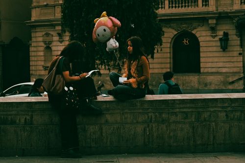 Free Two women sitting on a stone wall in a city holding a cartoon balloon. Stock Photo