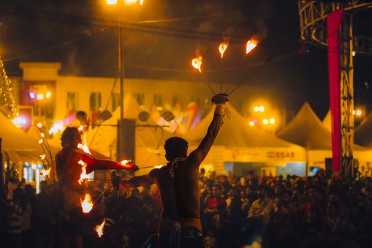 Crowd mesmerized by fire dancers under vibrant festival lights at night.