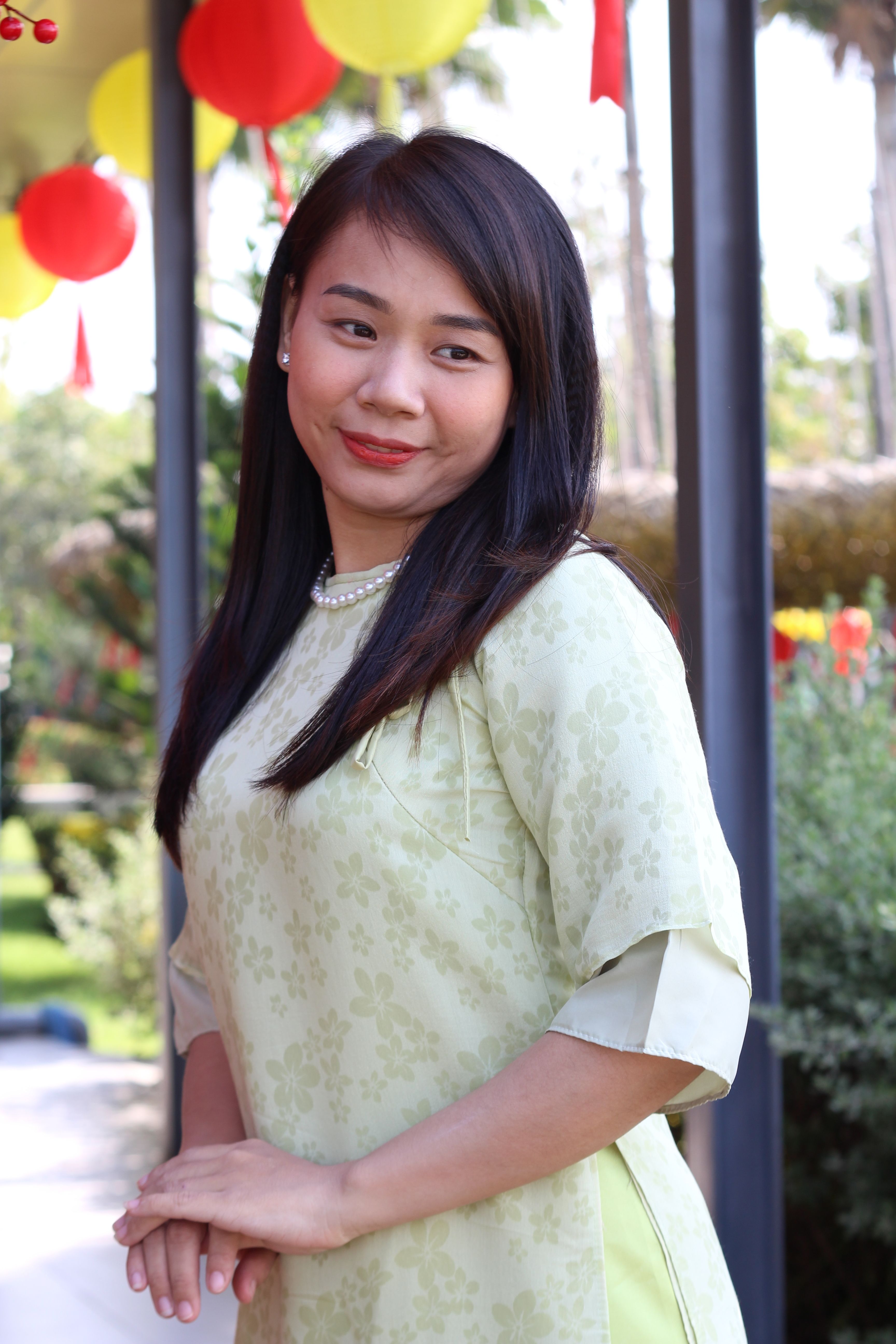 A woman in a green áo dài dress smiles outdoors with festive decorations.