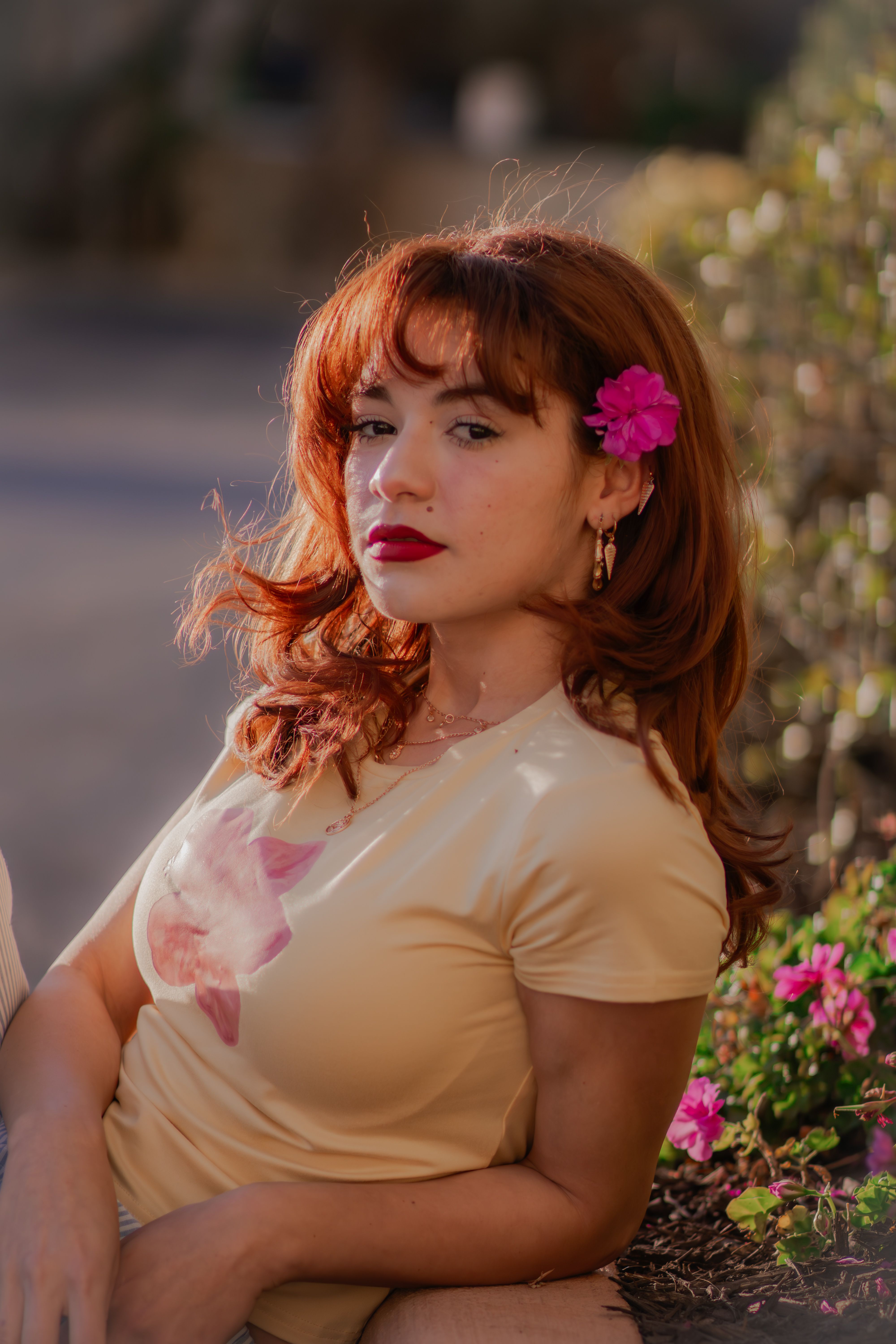 Elegant woman with red hair and pink flower in her hair lounging outdoors in soft light.