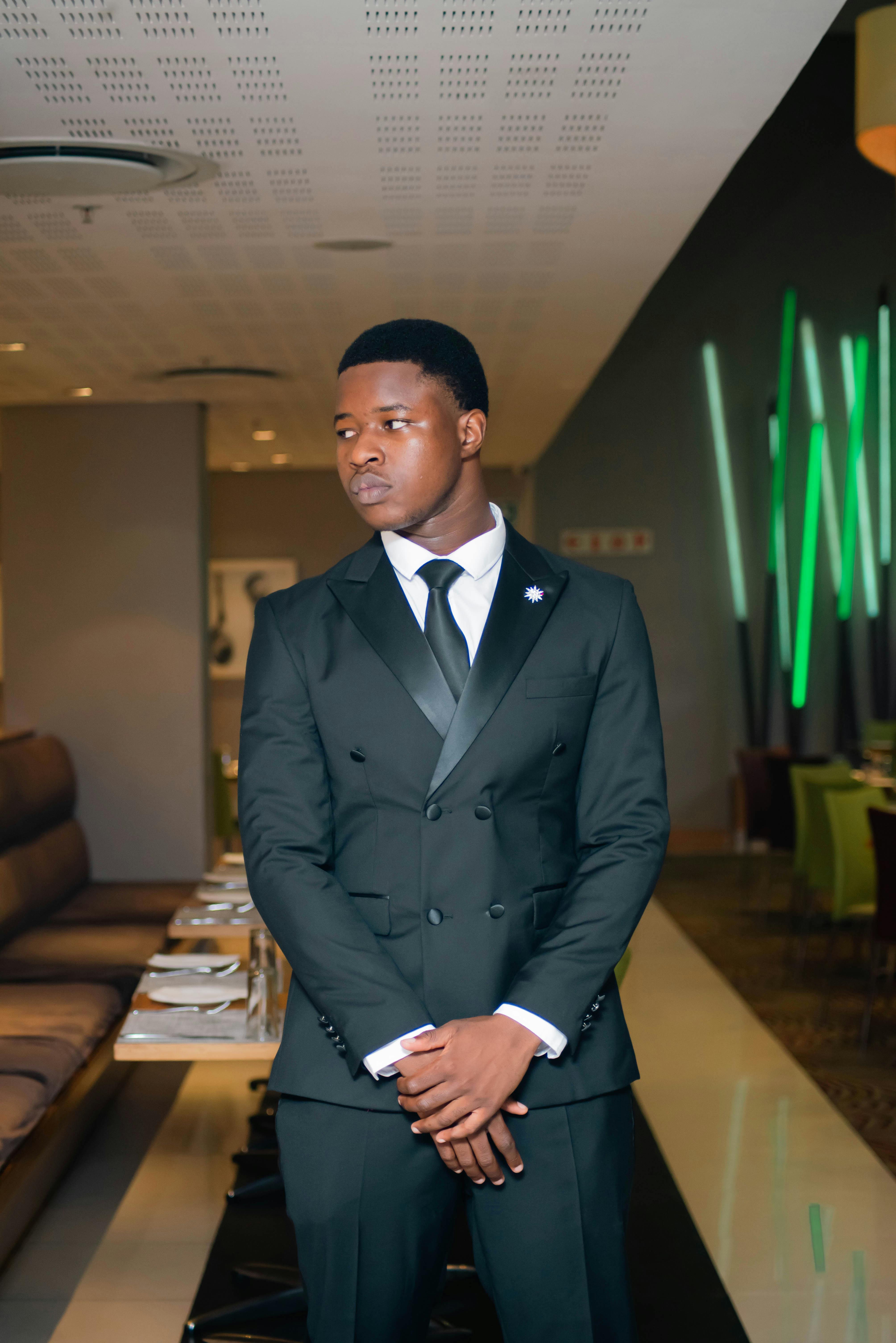 A stylish young man in a black suit stands poised in a modern restaurant setting.