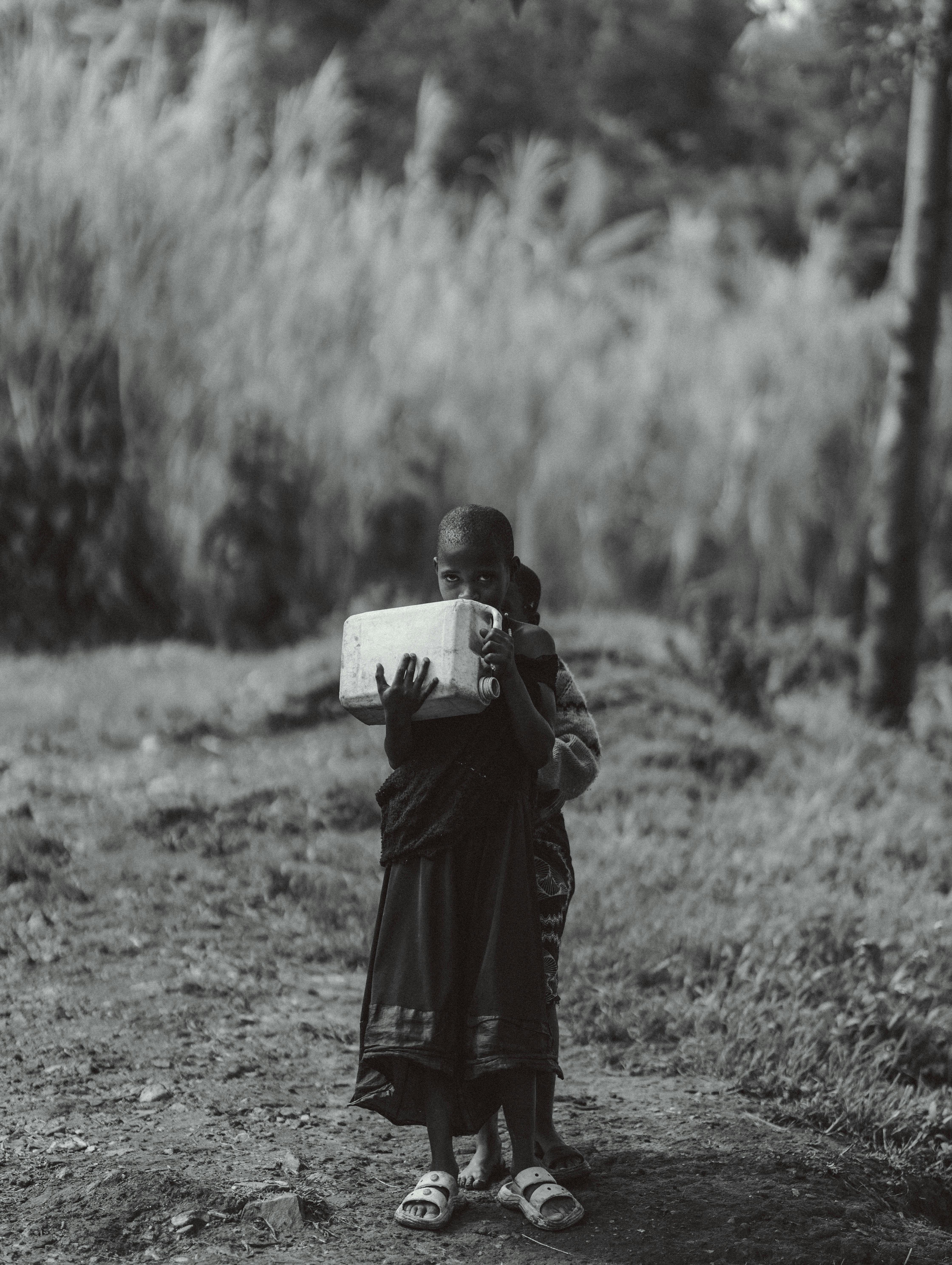 Black and white image of a child carrying a large water container on a rural path.