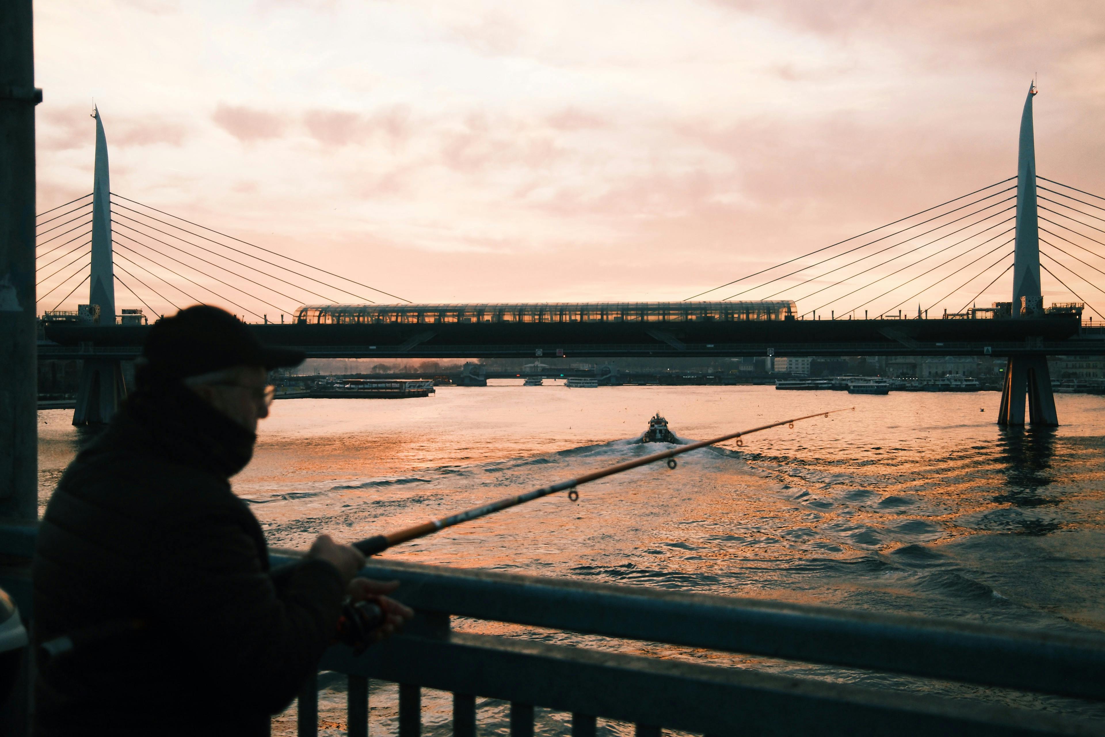 Silhouette of a fisherman against a scenic urban backdrop of a cable-stayed bridge at sunset.